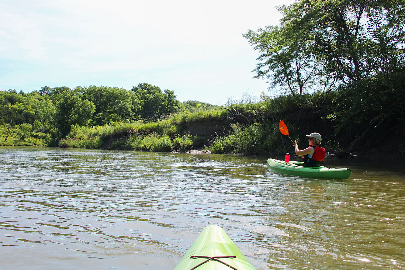 a girl kayaking on the a river with green grass and trees and blue sky on horizon