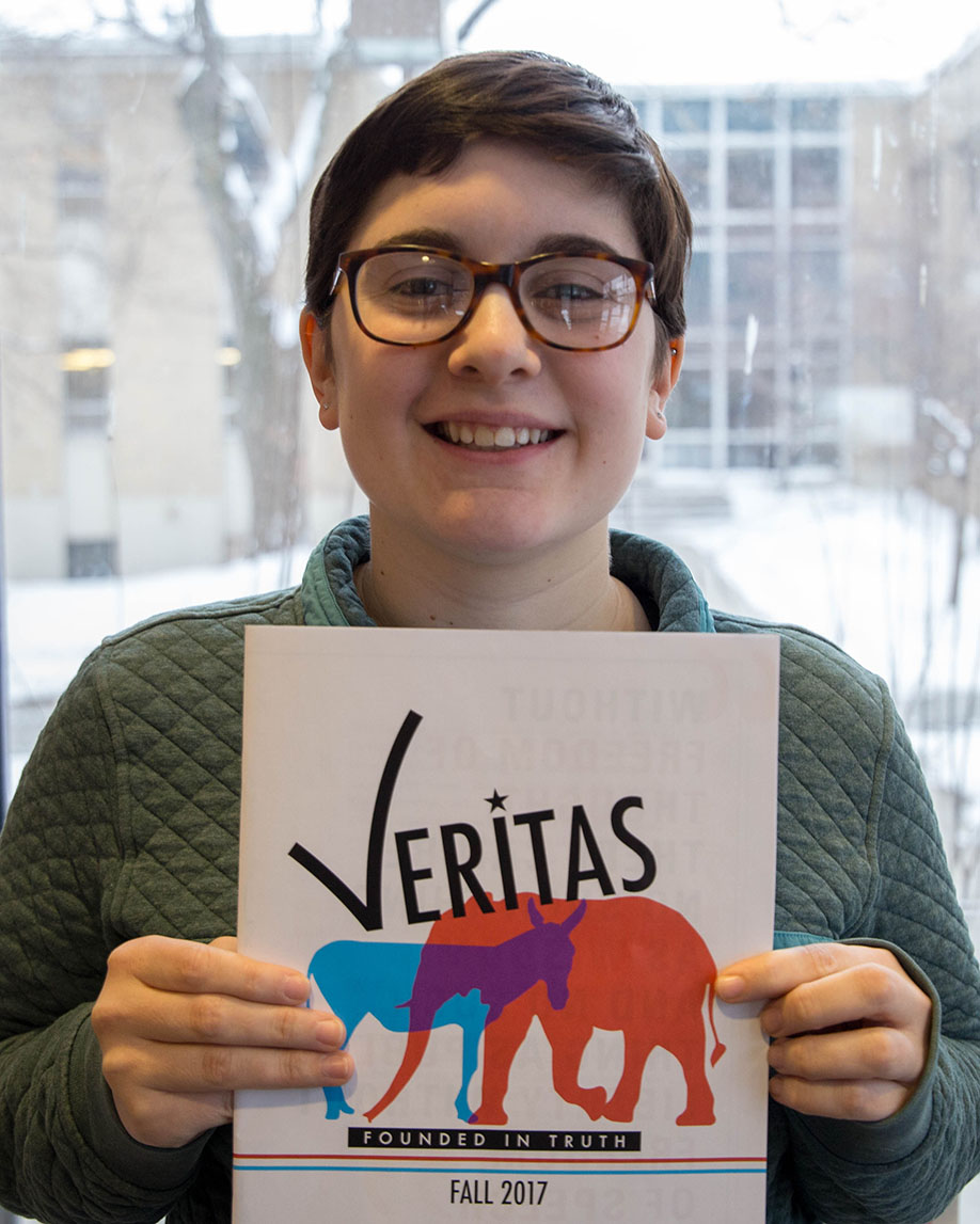 Courtney Carstens has glasses, short brown hair and is holding up a copy of Veritas with a donkey and elephant on the cover