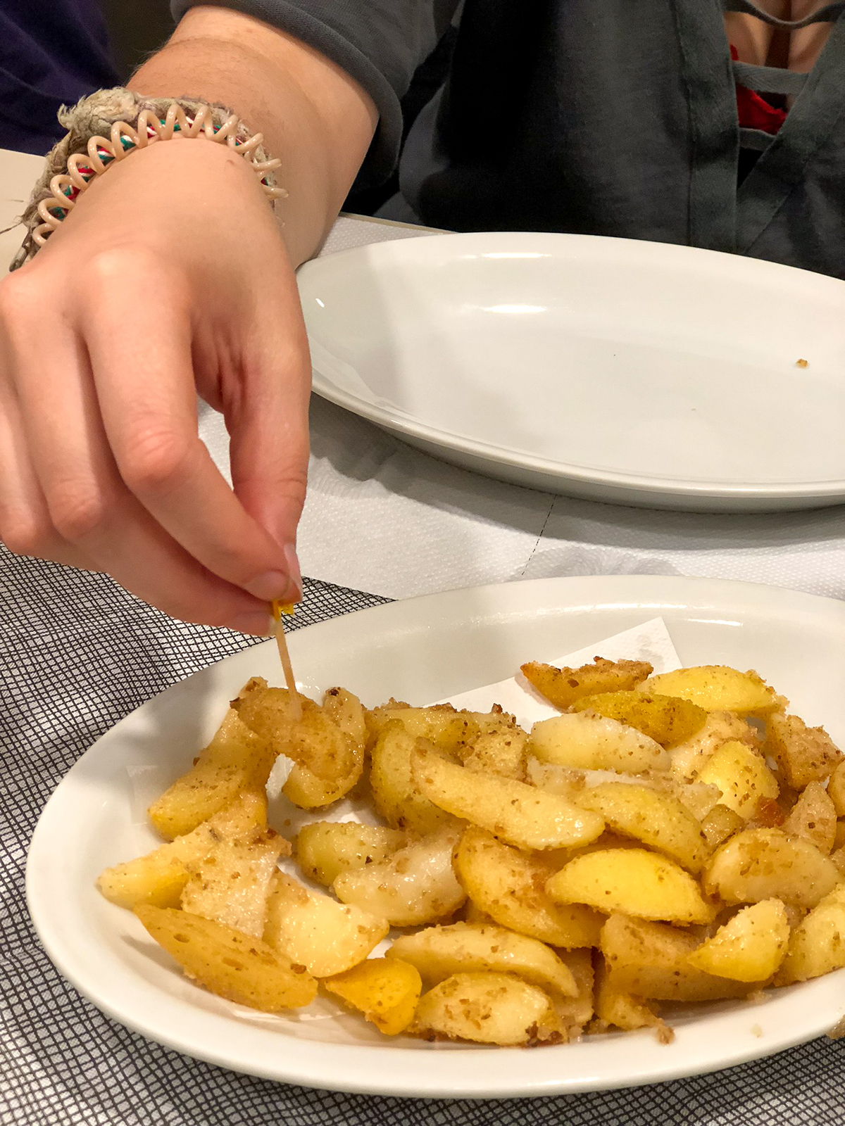 A hand reaches to pick up a small piece of potato from a white plate of potatoes.