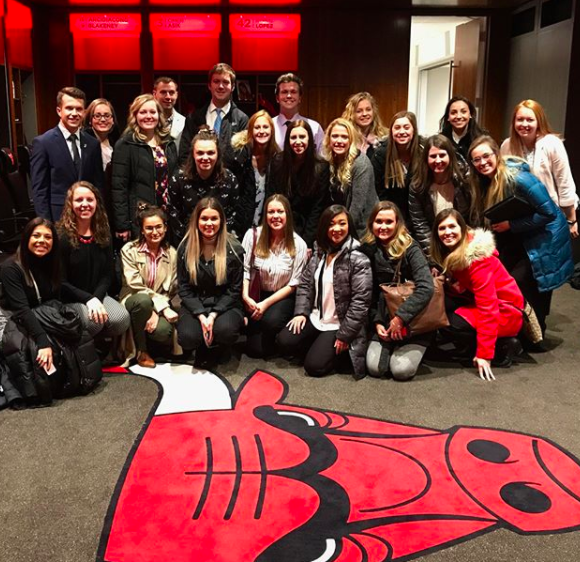 A group of students in winter coats huddle for a group photo with a big Chicago Bulls logo in front of them on the floor.