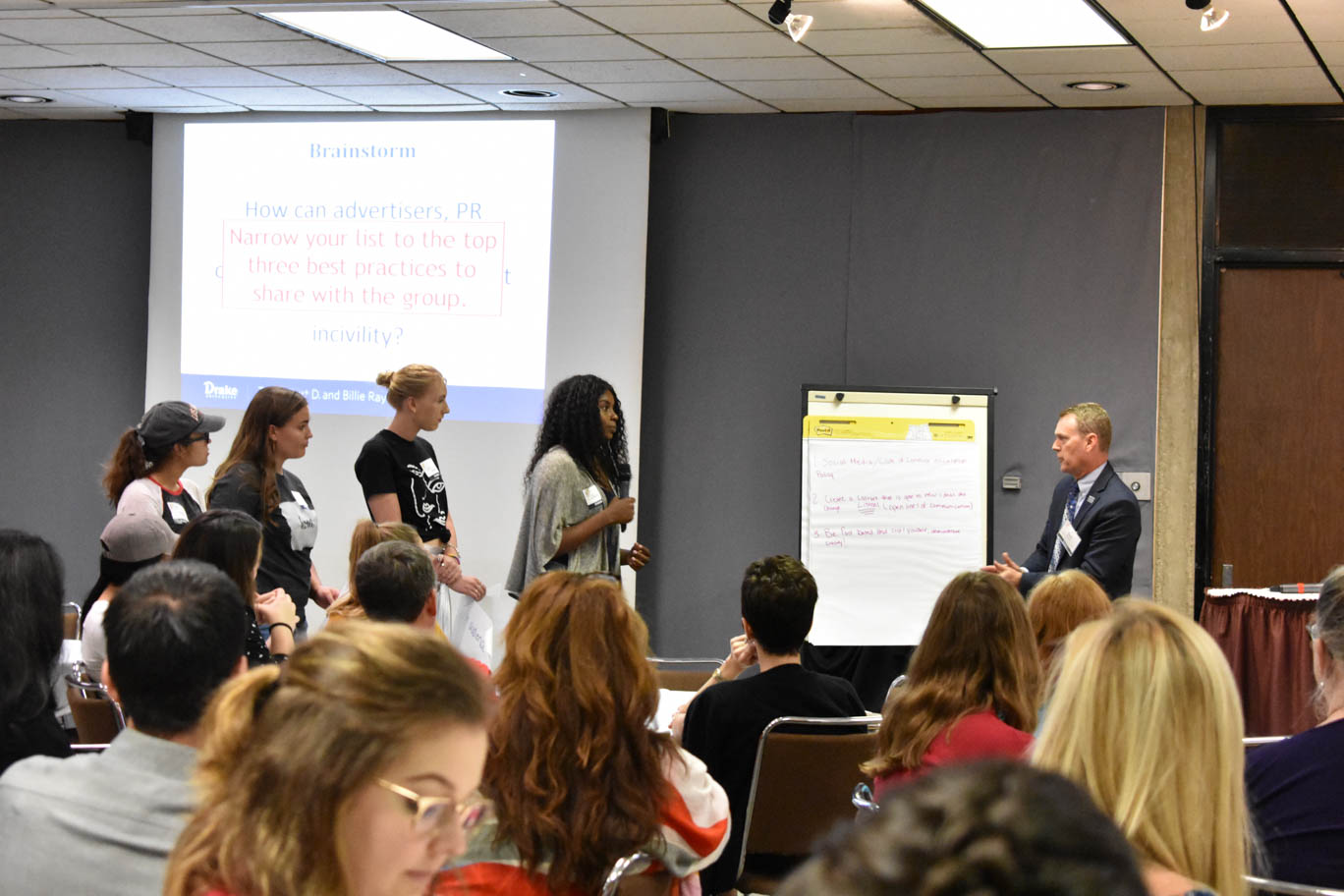 Studnets and a moderator standing in front of a white notepad in a conference room