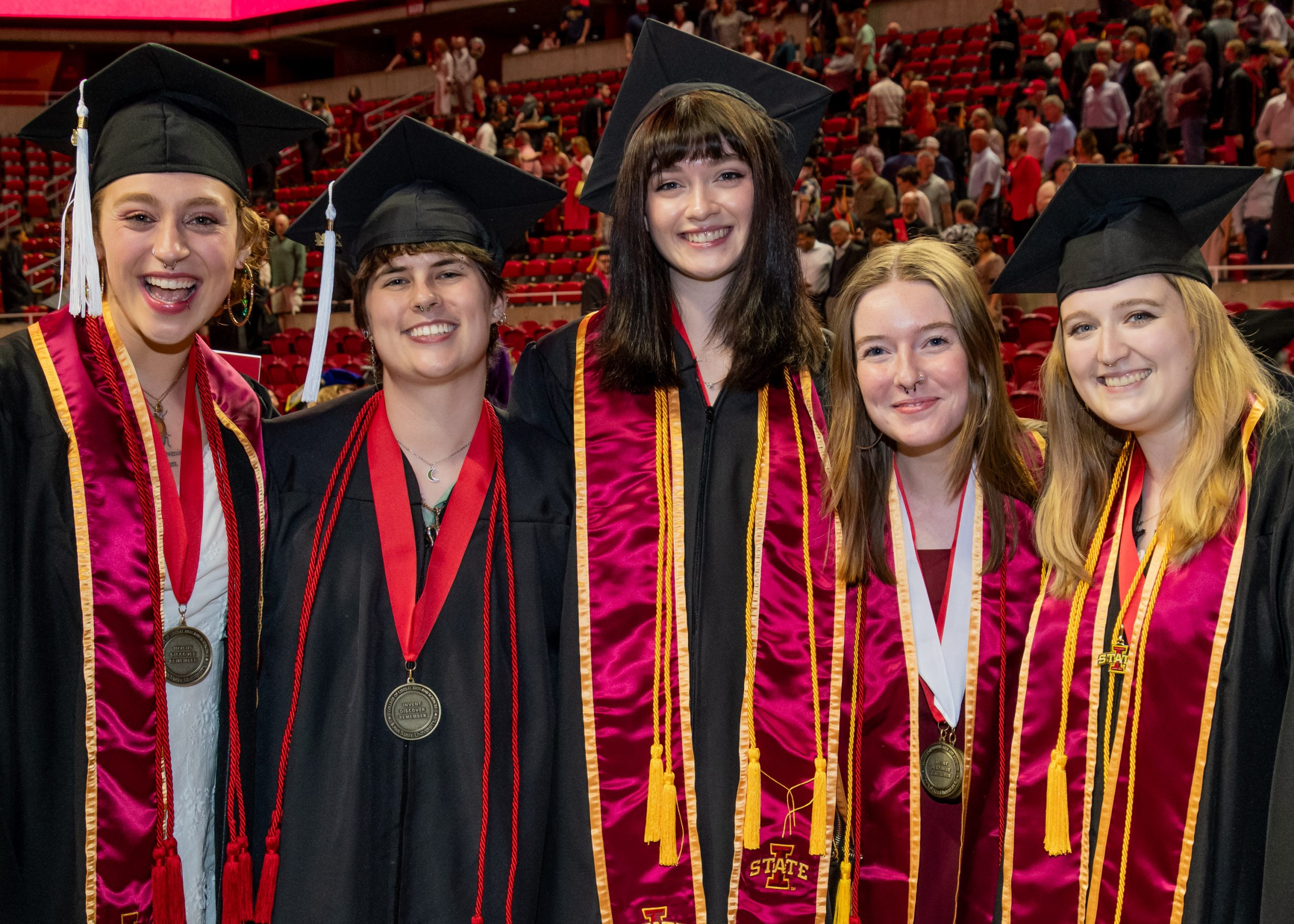 Five students in a row wearing regalia and LAS medallions smiling at the camera after the Spring 2024 LAS Convocation with audience members exiting in the background.