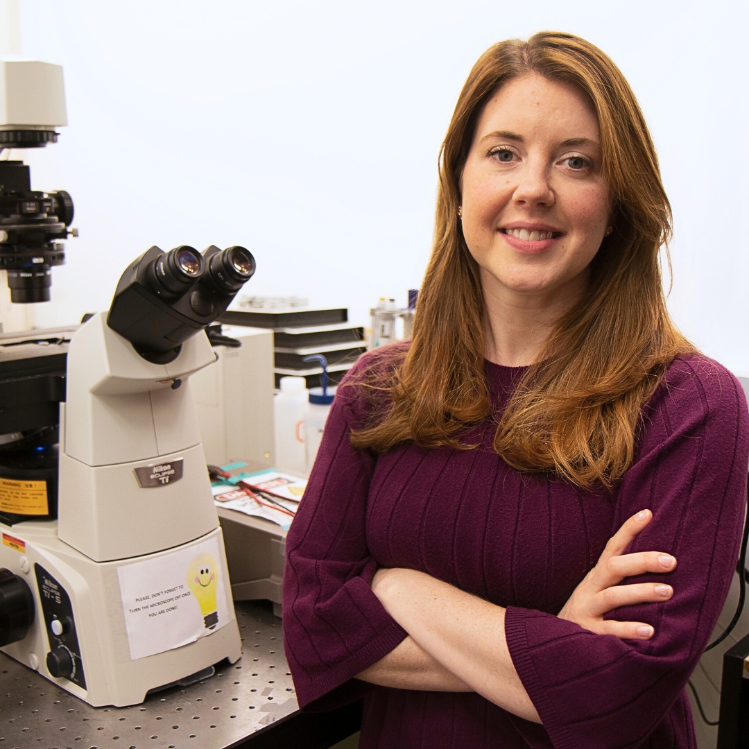 Robbyn Anand, associate professor in Chemistry, poses cross-armed wearing a purple-knitted sweater next to a microscope with a graphic of a lightbulb with a face attached.