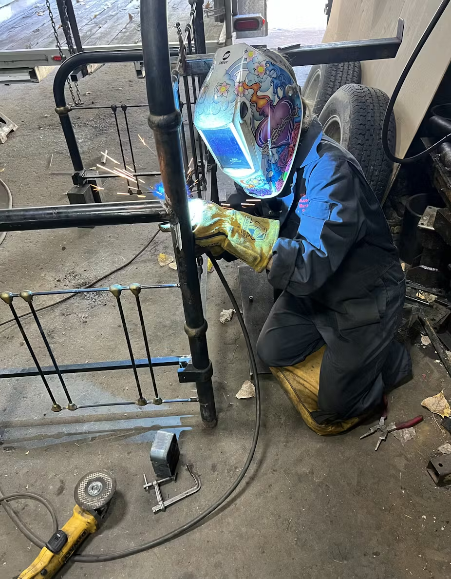 Jo Mackiewicz, professor in English, sporting a welders mask and protective gloves as she welds the headboard of a bed frame.