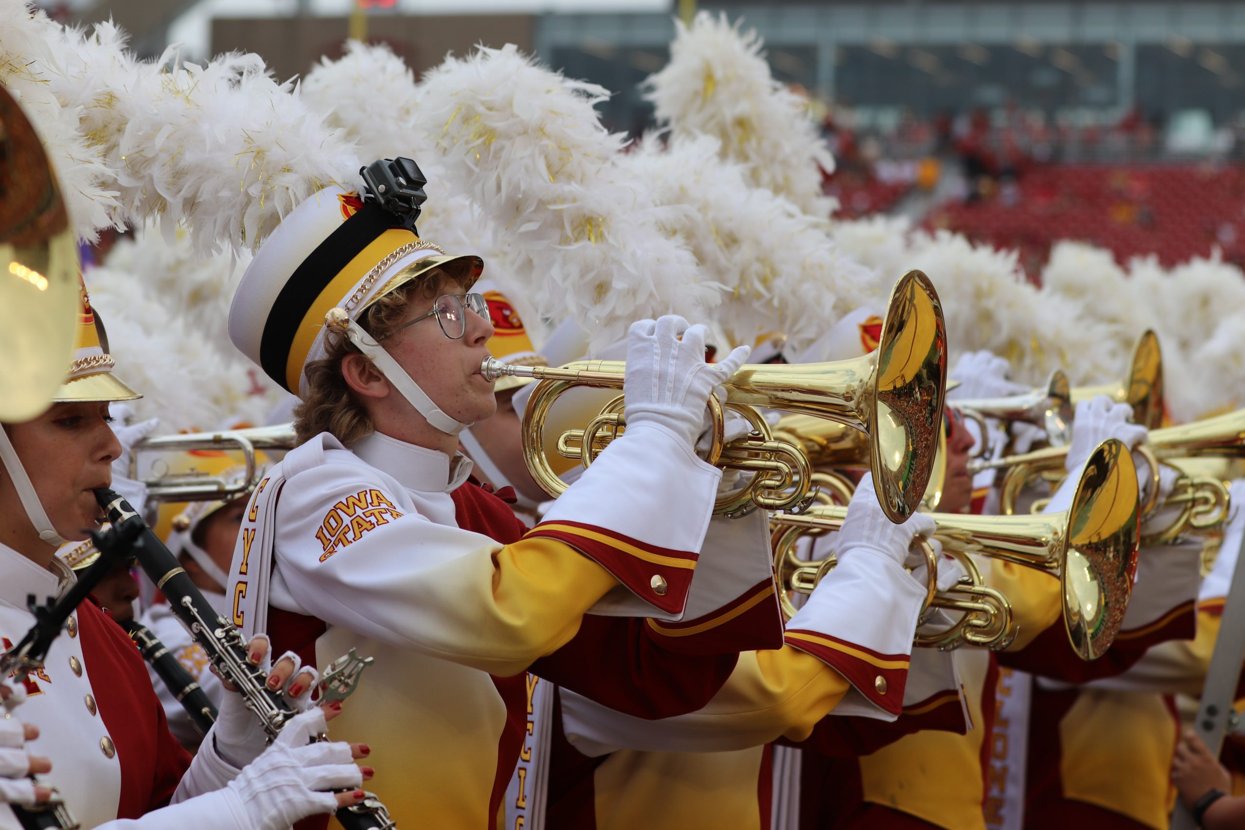 A group of Iowa State University Cyclone Football 'Varsity' Marching Band members performing. A trumpet player in the foreground, wearing a white and gold uniform with a feathered hat, plays their instrument with focus. Other band members and instruments, including clarinets and brass, are visible in the background, along with a blurred stadium setting.