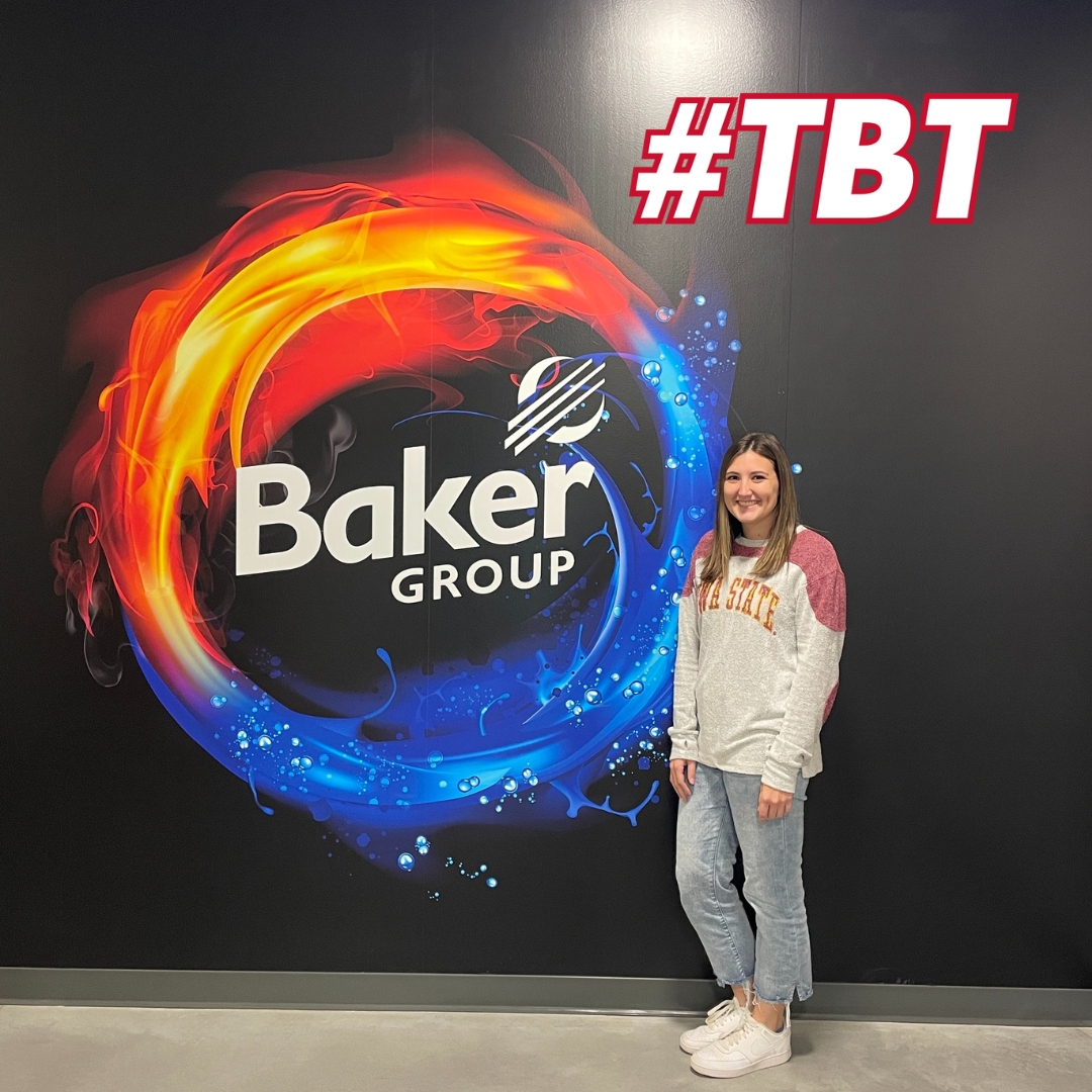 Brooke Schaben ('16 communication studies) stands in front of a large Baker Group company logo on a black wall. Schaben is wearing an Iowa State long-sleeve shirt and jeans.