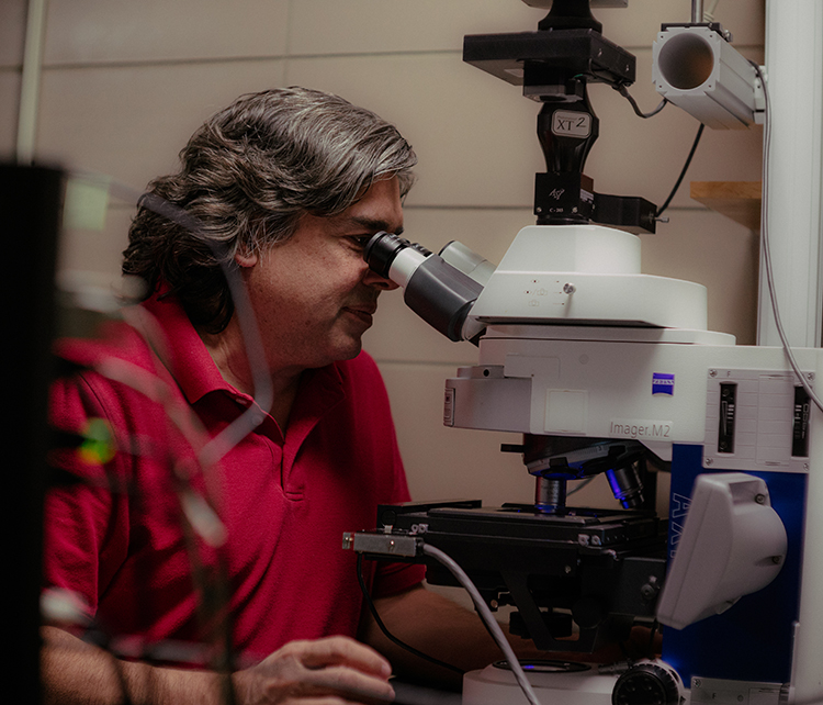 Mohan Gupta looking into a high-powered microscope in a laboratory. The microscope is equipped with various components, including cameras, lenses, and wires, indicating advanced scientific equipment.