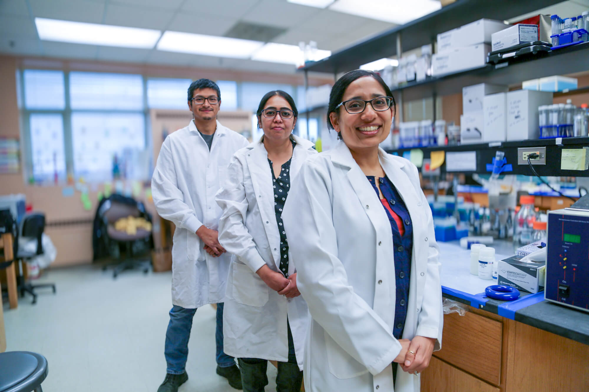 Three researchers in white lab coats standing in a laboratory. The person in front is smiling warmly, while the two in the background stand with their hands clasped. Shelves filled with laboratory equipment and supplies are visible in the background.