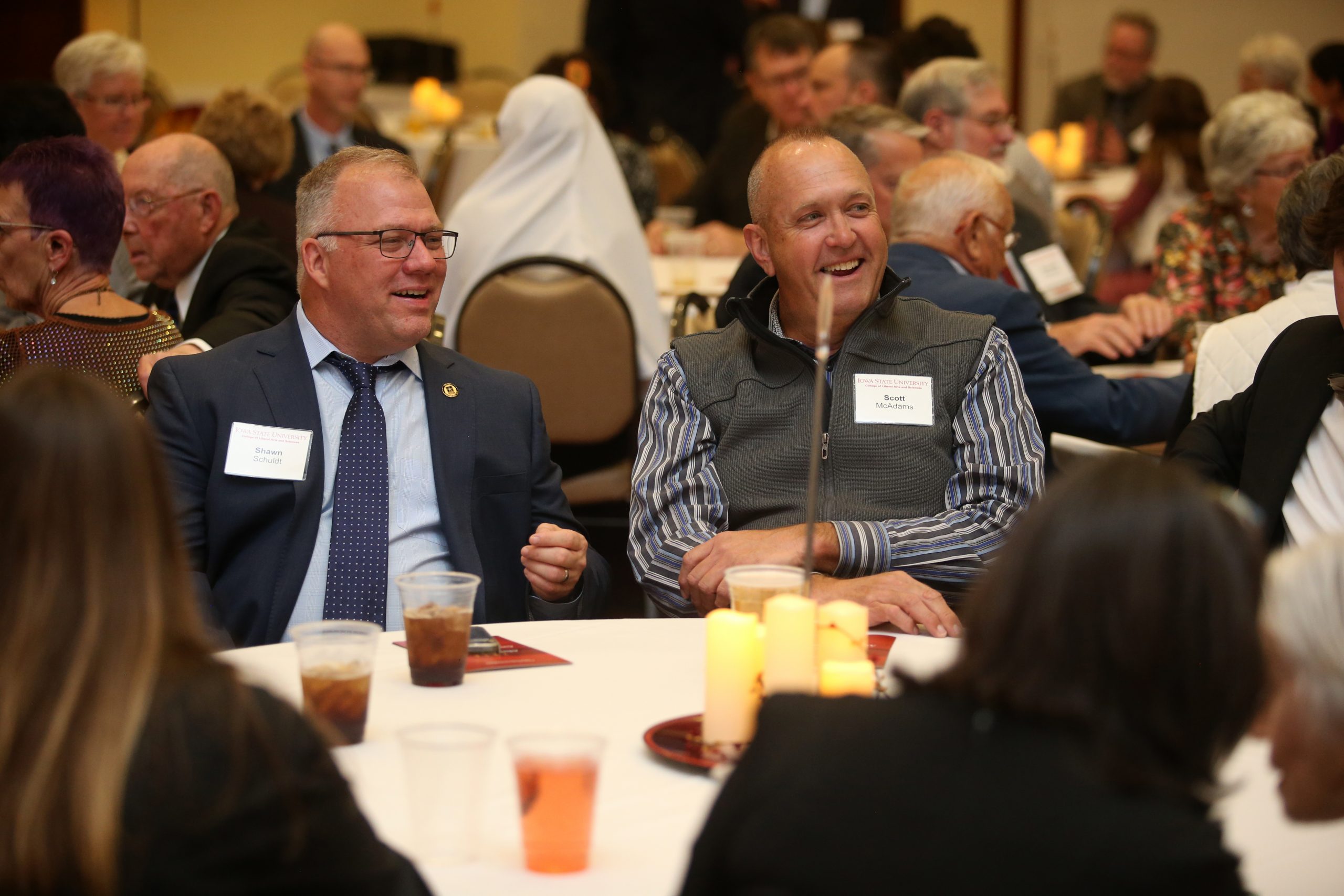 Two men are seated at a round table during a formal gathering. The man on the left, wearing glasses, a suit, and a tie, is smiling and appears engaged in conversation. His name tag reads 'Shawn Schuldt.' The man on the right, dressed in a striped shirt and a vest, is also smiling, with his name tag reading 'Scott McAdams.' The table is decorated with candles, and other attendees are visible in the background, seated and talking.