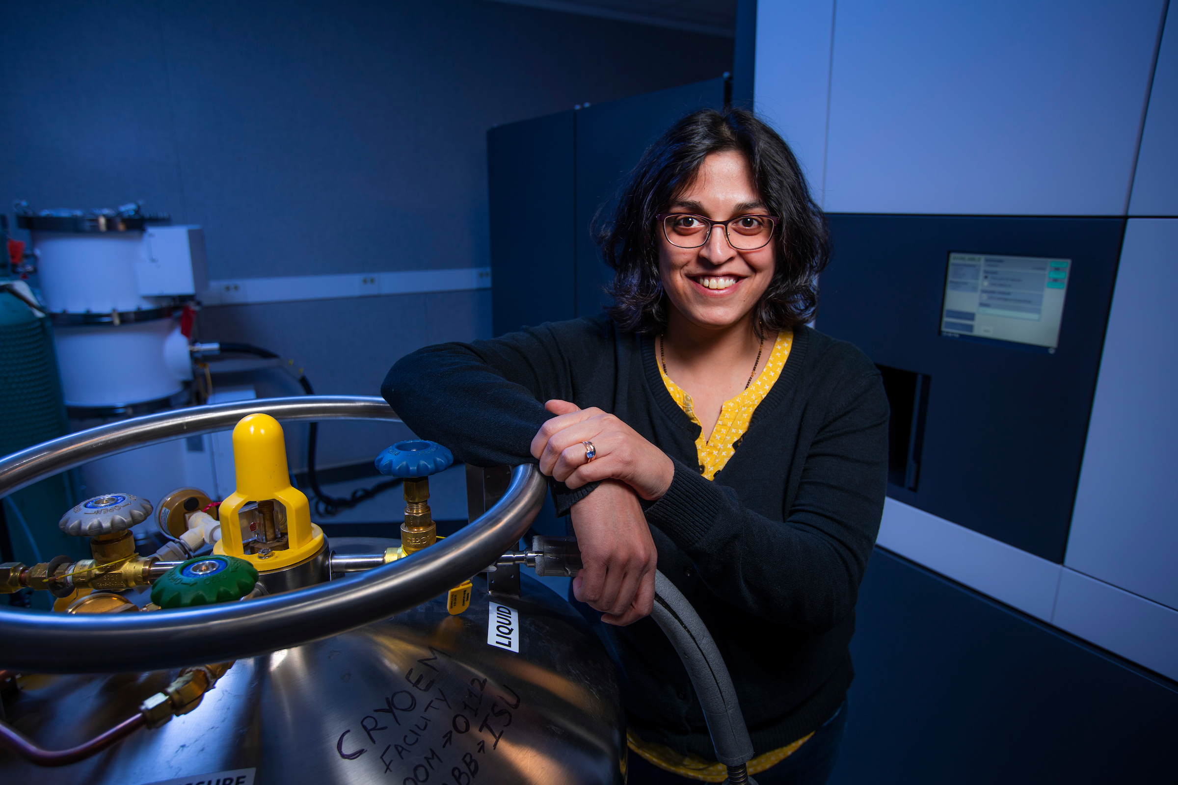 Dipali Sashital smiles confidently while standing in a laboratory. She leans on a cryogenic liquid storage tank with colorful valves and labeled 'CRYOEM' in black marker. She wears a black cardigan over a yellow patterned top, with scientific equipment and monitors visible in the background.