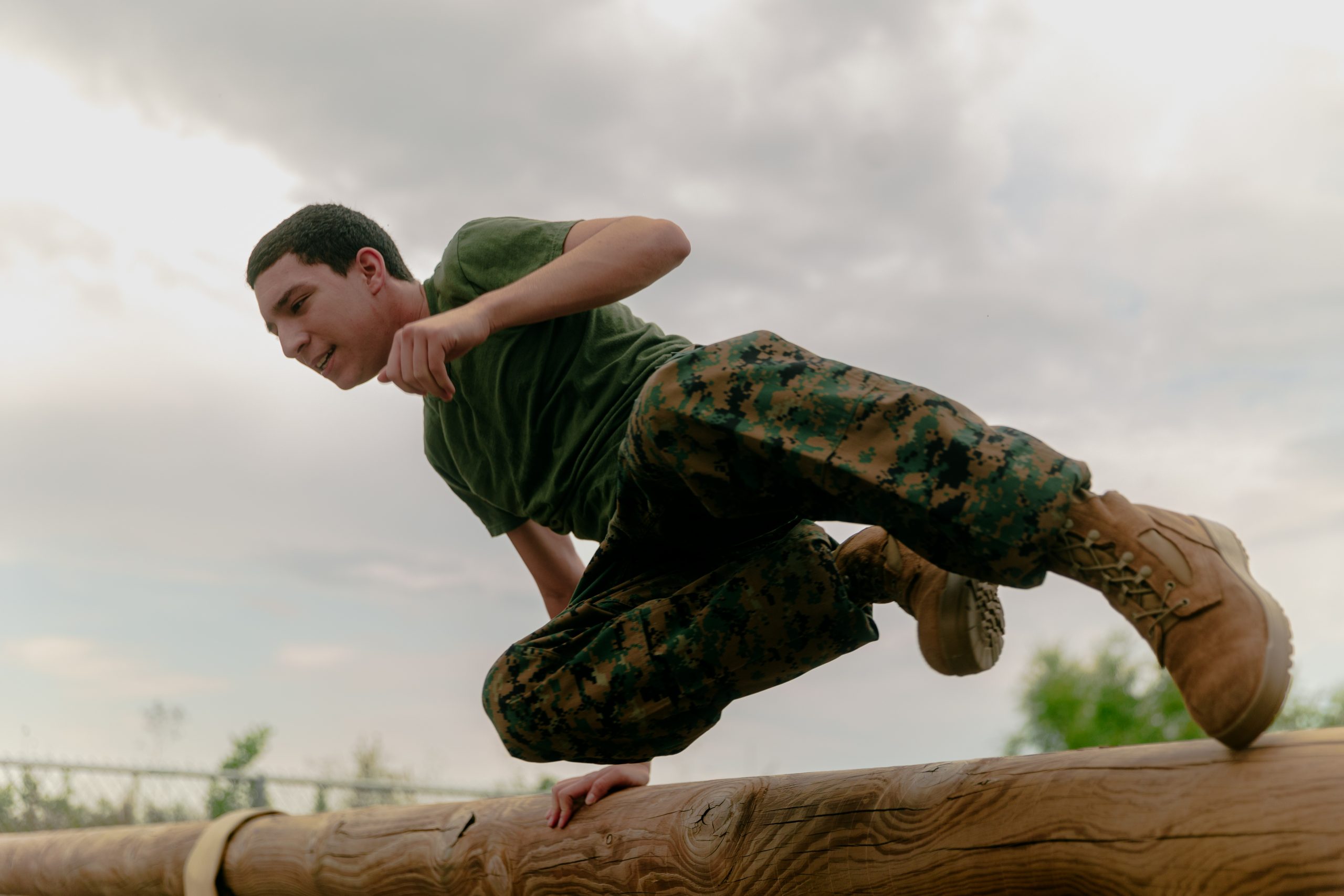 A young man wearing a green military-style T-shirt and camouflage pants is captured mid-action as he leaps over a wooden log obstacle. He is using one hand to support himself while swinging his legs over, with a determined expression on his face. The background consists of a cloudy sky and some greenery.