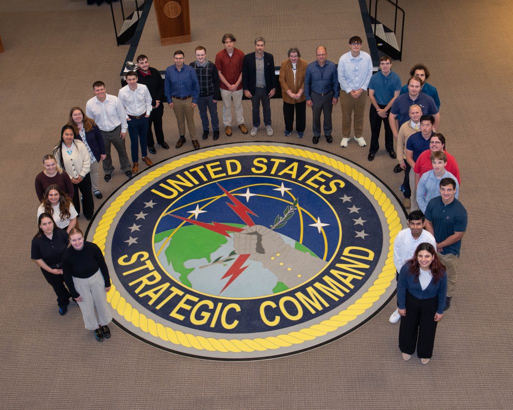 A group of approximately 30 people stands in a semi-circle around the United States Strategic Command emblem on the floor. The emblem features a clenched fist holding lightning bolts over a globe, surrounded by stars and the text "United States Strategic Command." The group consists of men and women of diverse ages and is photographed from above in a large room with beige carpeting. A podium is visible in the background.