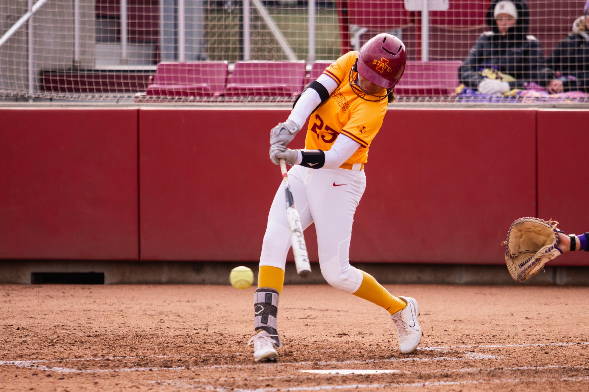 Angelina Allen wearing a yellow and white Iowa State University uniform with the number 23 is captured mid-swing, making contact with the ball. She is wearing a cardinal-colored helmet with the Iowa State logo, gray batting gloves, and a black arm sleeve. Her left leg has a protective brace, and she is wearing white cleats with gold socks. In the background, a catcher’s gloved hand reaches out, and spectators in warm clothing sit behind a protective net in the stands. The setting is a softball field with a dirt infield and red padded walls at the Cyclones Sports Complex.