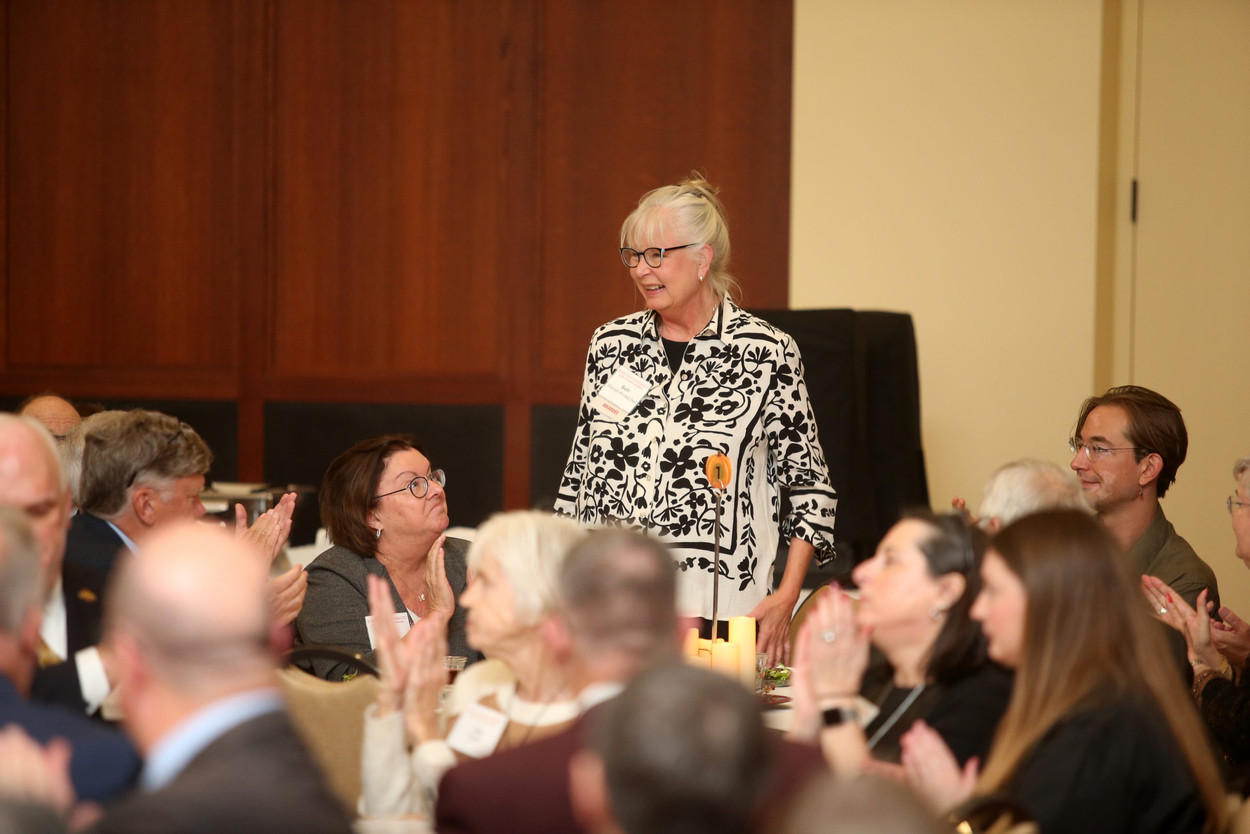 An Alumni Award recipient stands while others sit and applaud at the Alumni Awards event. She wears glasses and a patterned blouse.