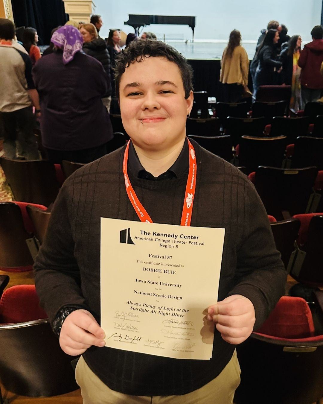 Bobbie Buie, senior in performing arts, smiling while holding an award certificate for National Scenic Design at the Kennedy Center American College Theatre Festival, wearing a dark sweater and a red lanyard. Audience visible in background.