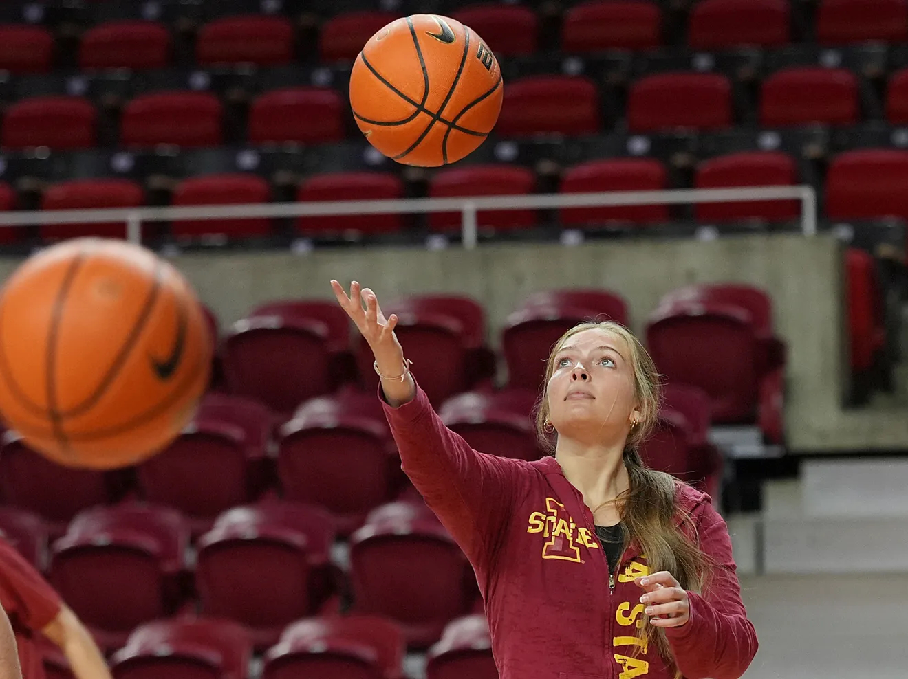 A college-aged Elise Lambert with long blonde hair pulled back, wearing a maroon Iowa State jacket, is on a basketball court practicing. She is reaching out with one hand as a basketball hovers mid-air in front of her. Another basketball is in the foreground, slightly out of focus. The background features empty maroon-colored stadium seats.