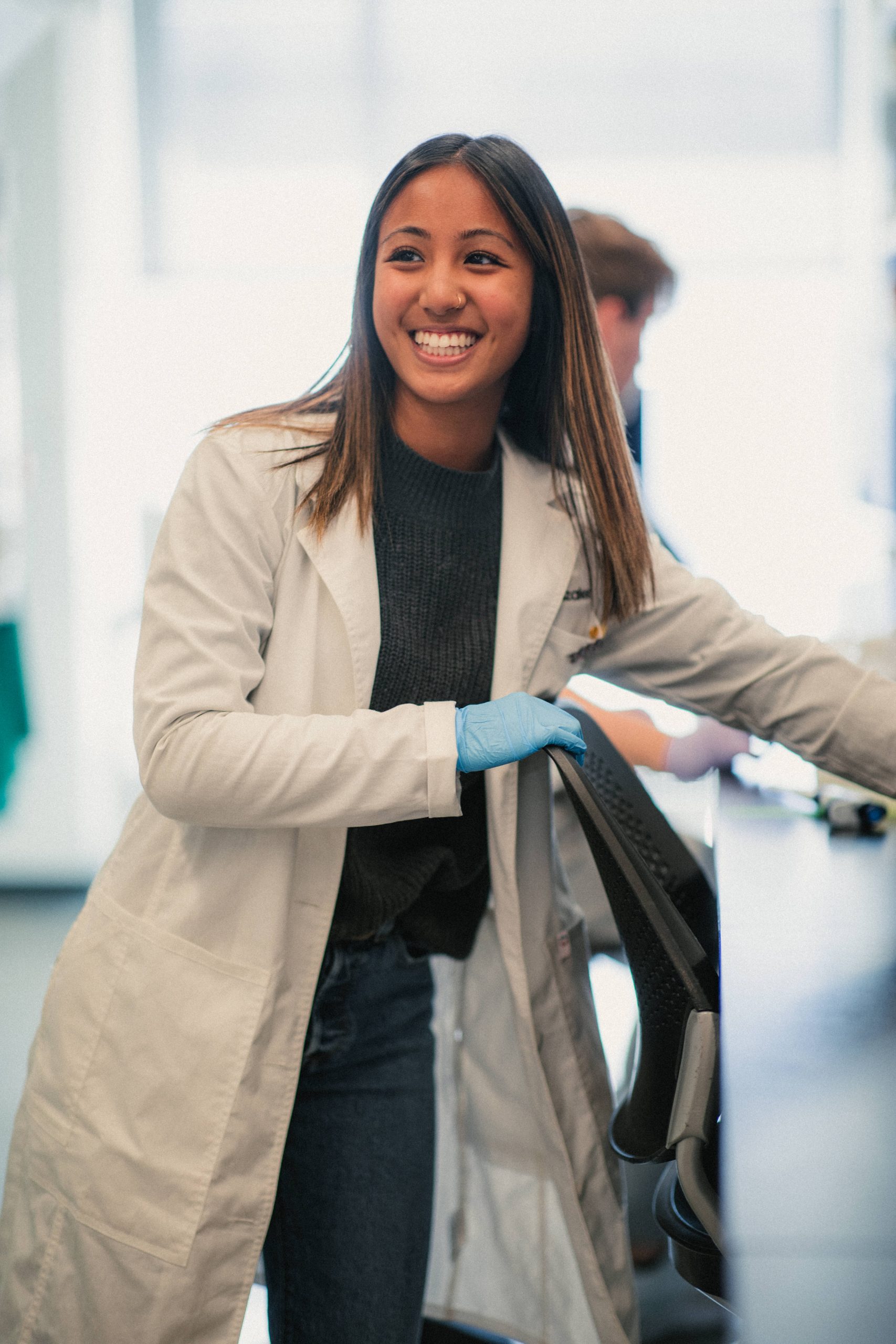 Caitlin Gonzales, wearing a white lab coat and blue gloves, smiles brightly while leaning on a chair in a laboratory. She appears engaged and confident, with a relaxed posture. In the background, another person is working at a lab bench, adding to the active research environment.