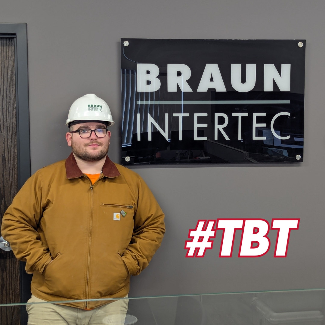 Canaan Mills wearing a white hard hat with the "Braun Intertec" logo, glasses, and a brown work jacket stands with his hands in his pockets in front of a black sign that reads "BRAUN INTERTEC" on a gray wall. He has a neutral expression. The reflection of an office setting is visible in the sign. A bold red and white "#TBT" text is overlaid on the image.