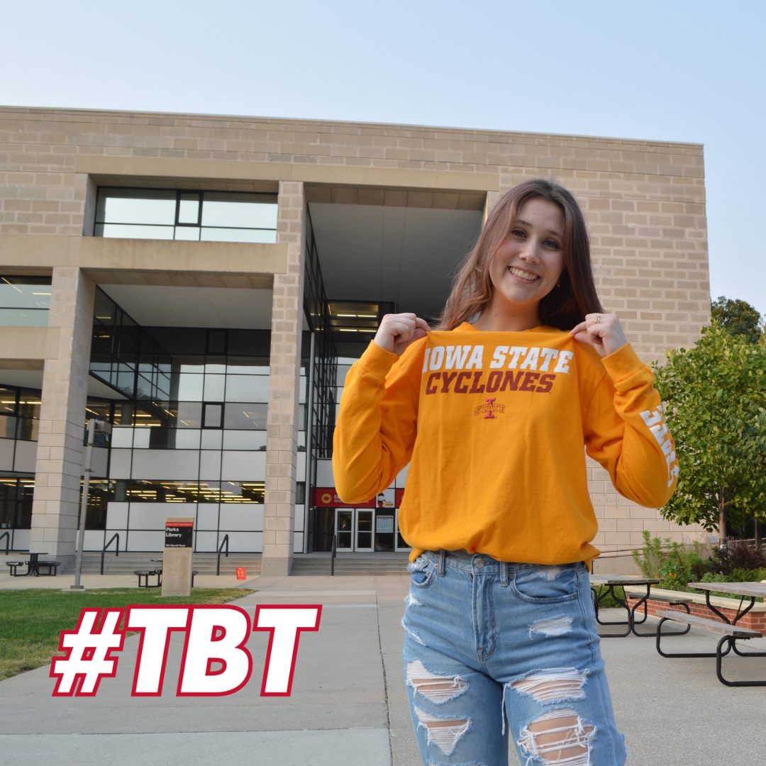 Devin Erb smiles while standing in front of Parks Library on the Iowa State University campus. She is wearing a bright yellow long-sleeve shirt with 'Iowa State Cyclones' written across the front and ripped light blue jeans. She holds the collar of her shirt with both hands, showing off the text. The hashtag '#TBT' in bold red and white text is placed in the bottom left corner of the image. Outdoor seating and greenery are visible in the background.