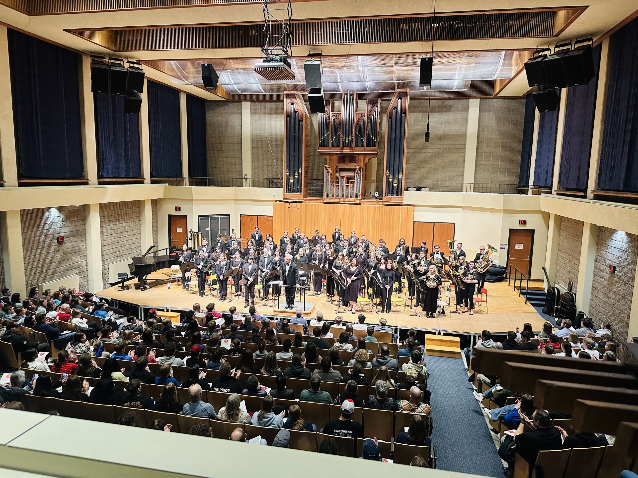 A large concert band performs on stage in a spacious auditorium with an audience watching. The musicians, dressed in formal black attire, stand and hold their instruments while the conductor speaks to the audience. The stage features a wooden backdrop with a large pipe organ in the center. The auditorium is well-lit, with tiered seating filled with spectators. Some audience members are clapping, while others hold programs. The setting has a modern architectural design with high ceilings, large speakers, and acoustic panels.