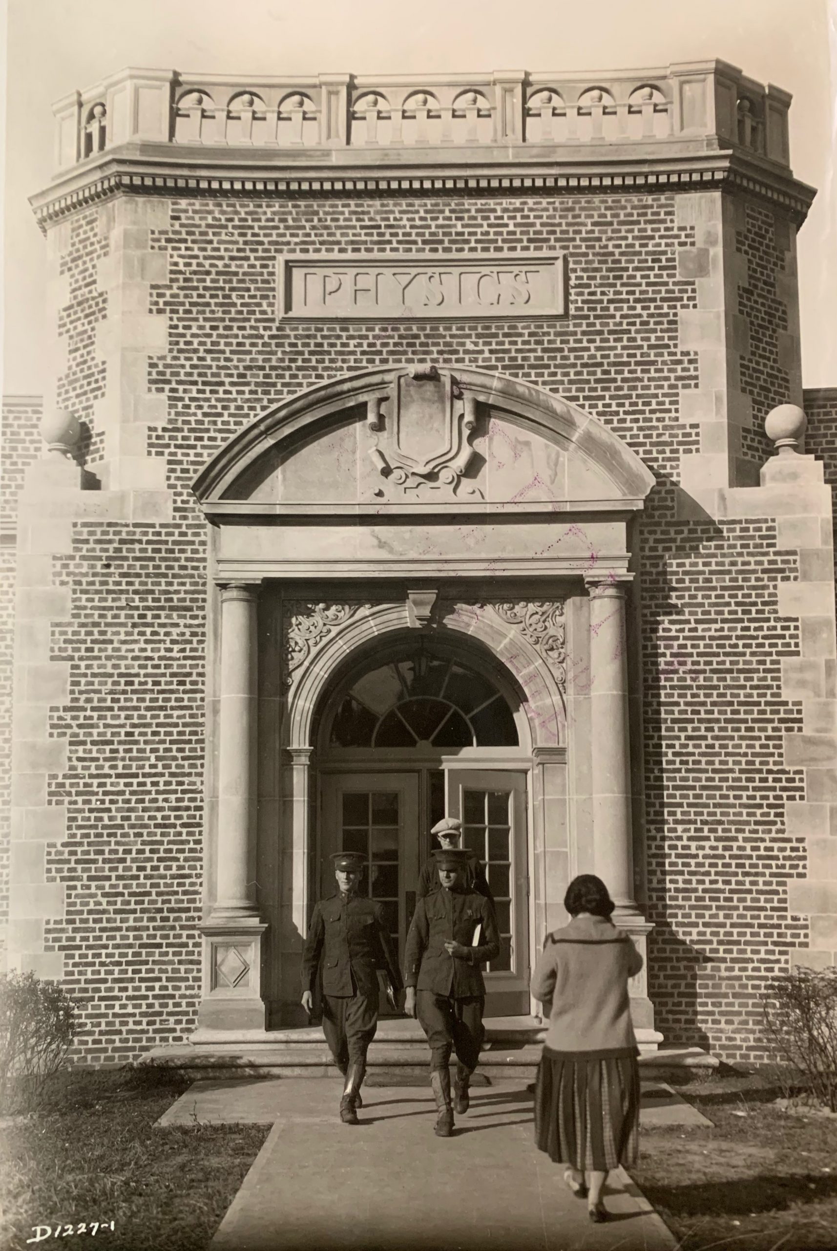 A black-and-white photograph from 1931 shows the exterior of a brick and stone physics building with an arched entrance. Above the entrance, a stone plaque reads "PHYSICS." Three men in military-style uniforms exit the building, wearing knee-high boots and caps, while a woman in a long skirt and a sweater walks toward the entrance. The photograph captures a moment of daily activity, with strong shadows suggesting it was taken on a sunny day. The architectural details include decorative stonework around the door and a balustrade along the roofline.