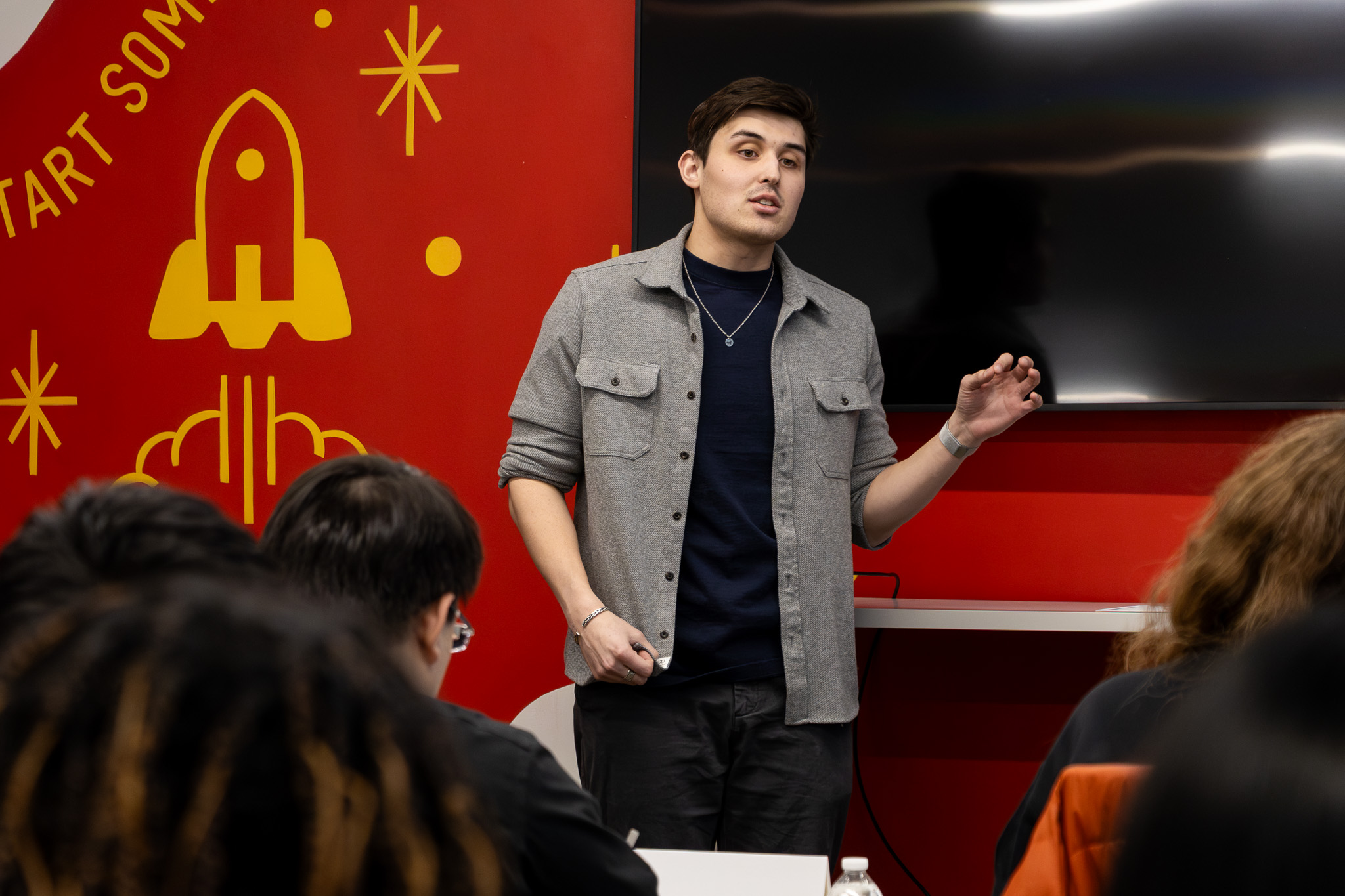 A young man with light skin, short blond hair, and a plaid blue-and-yellow button-up shirt is giving a presentation. He holds up a printed spectrogram with a purple-to-orange gradient while gesturing with his other hand. A large screen is behind him, and a red-and-white wall design is visible in the background. Audience members are partially visible in the foreground.