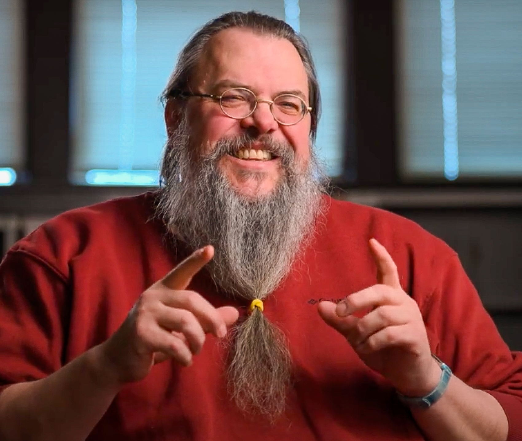 Steve Butler with glasses, wearing a red sweater, smiles warmly while gesturing with both hands. He has a long gray beard tied with a yellow band and is sitting in a well-lit room with windows in the background.