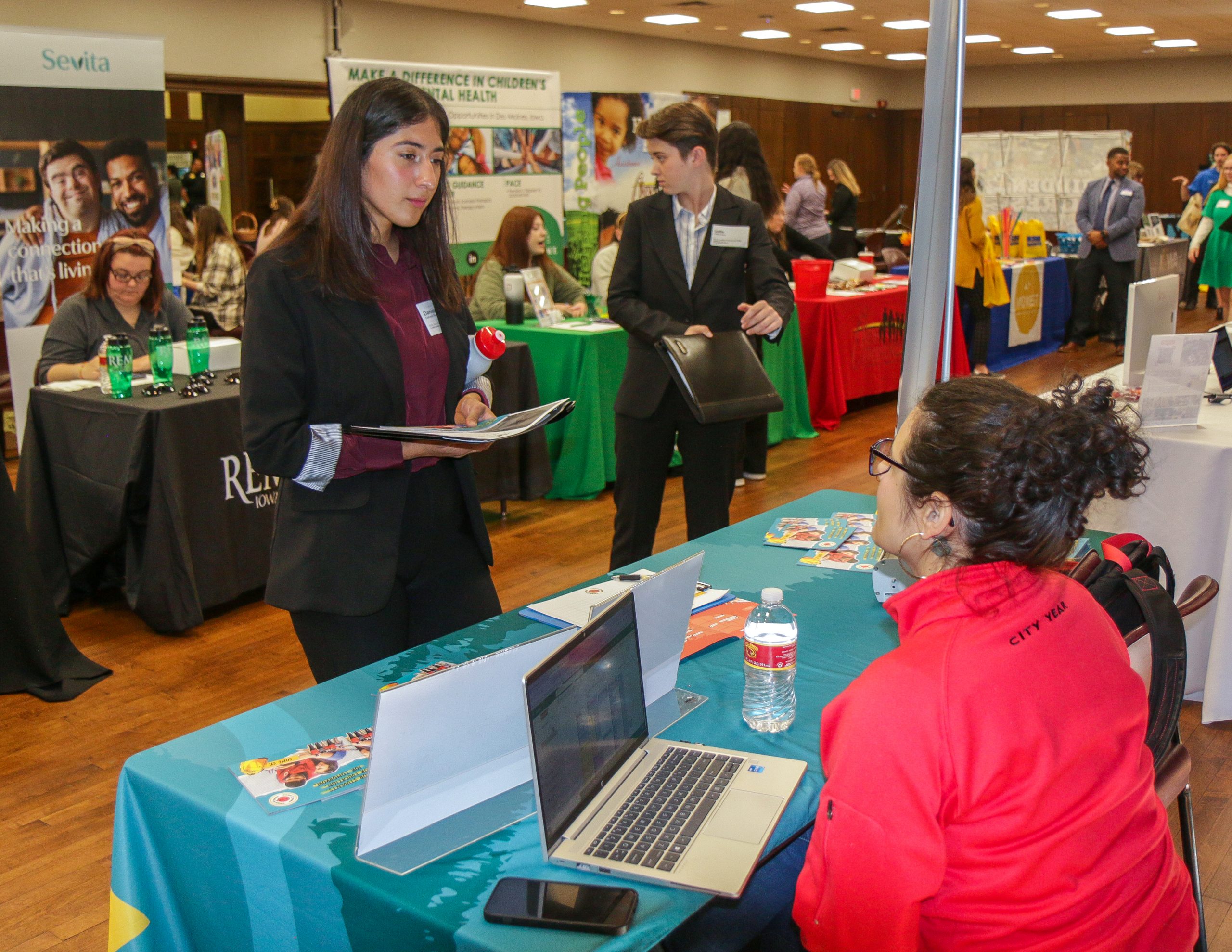 A career fair is taking place in a large room with tables covered in colorful tablecloths, representing different organizations. In the foreground, a woman with dark hair tied back, wearing a red "City Year" jacket, sits behind a table with a laptop, brochures, and a water bottle. She is engaged in conversation with a young woman standing in front of her, who is wearing a black blazer over a burgundy shirt, holding a portfolio and a red water bottle. Another individual in a black suit stands nearby, also holding a portfolio. In the background, multiple booths with banners and representatives are visible, along with other attendees dressed in business attire, networking and gathering information. The atmosphere is professional and bustling.