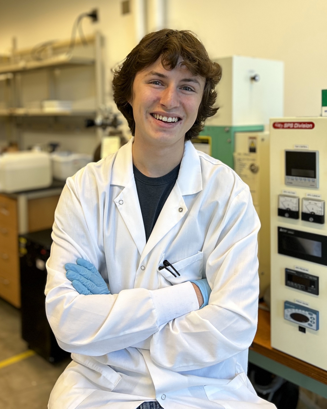 The image shows a person wearing a white lab coat and blue gloves, seated in a laboratory setting. They have brown hair and are smiling. The person is leaning slightly back with their arms crossed. In the background, there are shelves with various laboratory equipment and machinery. The walls are a light cream color, and the room appears well-lit. To the right of the person, there is a beige-colored cabinet with several dials and a label.