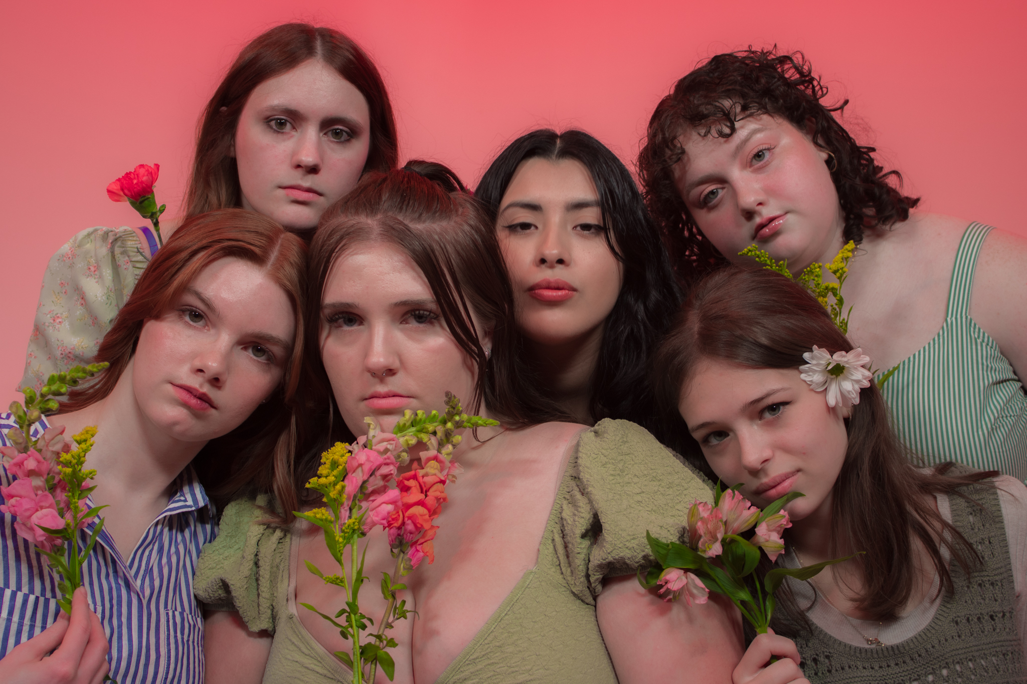 Six young women closely gathered against a gradient pink and red background, each holding or wearing fresh flowers. They have serious, serene expressions, and their outfits are soft and earthy-toned, complementing the natural floral theme.