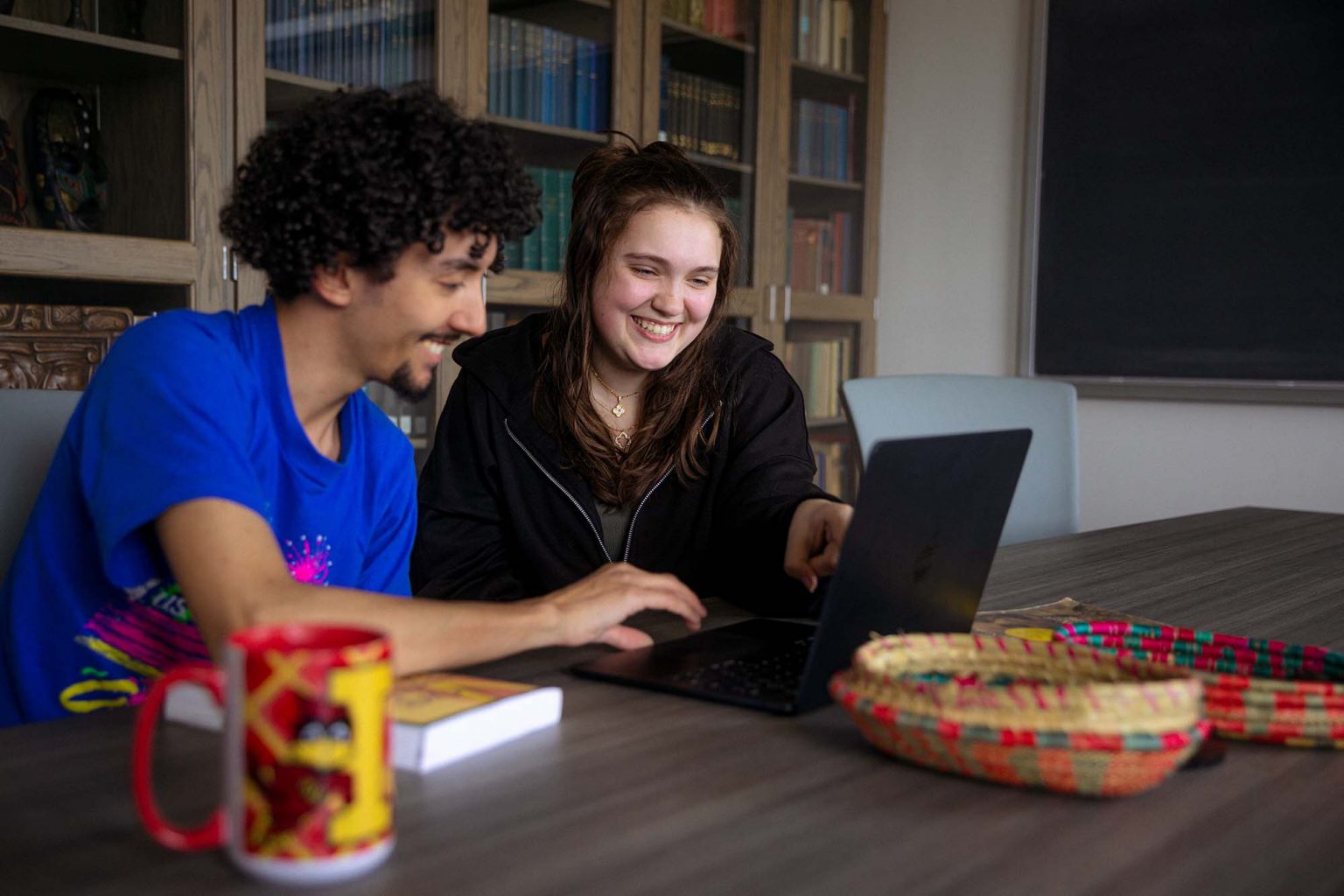 Two college students sit at a table in a library or classroom, smiling and looking at a laptop screen. One student, wearing a bright blue T-shirt, types on the keyboard, while the other, in a black hoodie, points at the screen. In the foreground are a colorful woven basket, a red and yellow mug, and a book. Shelves filled with books and a chalkboard are visible in the background.