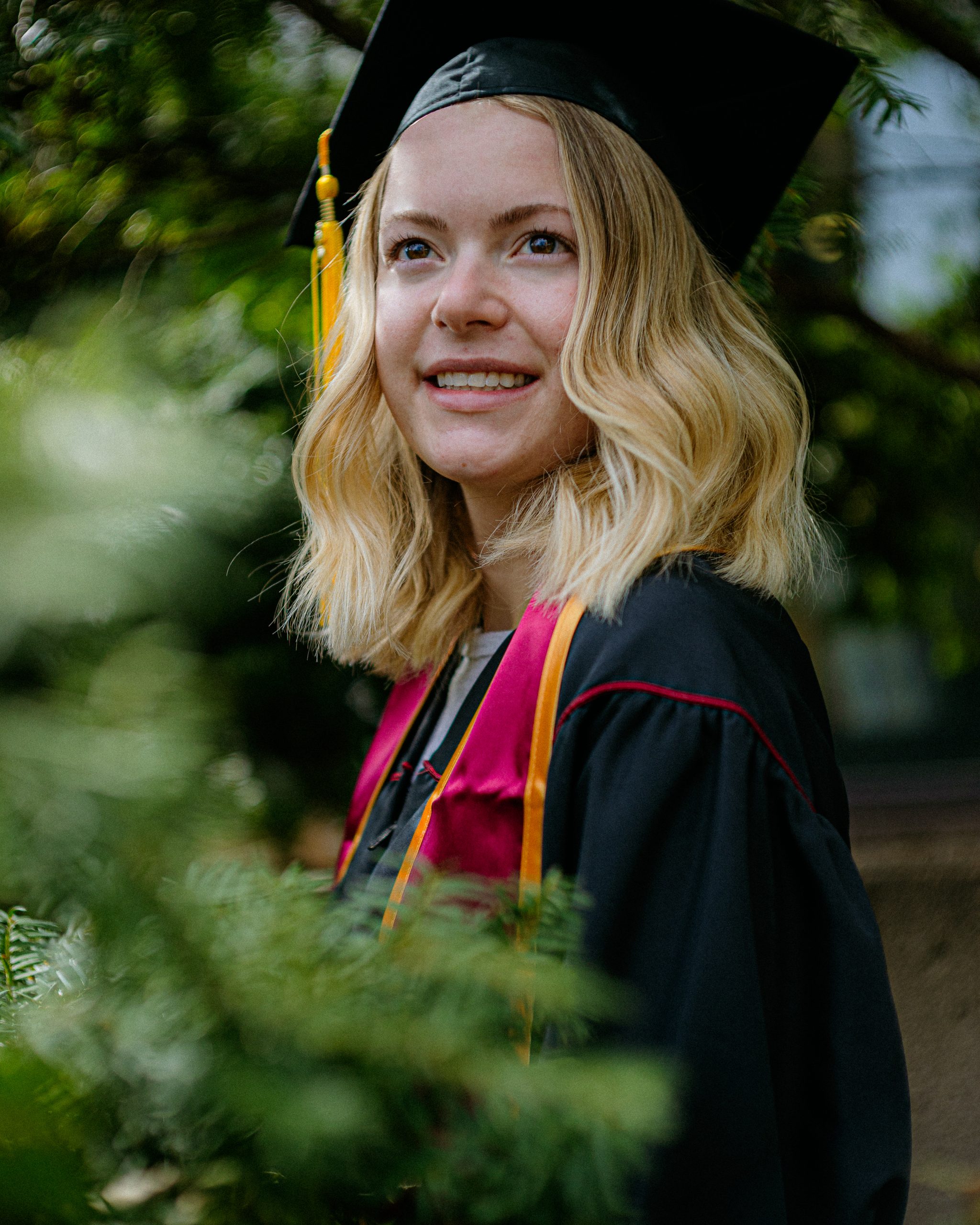A young woman in a black graduation cap and gown with gold and cardinal accents stands outdoors, smiling and looking into the distance. She is surrounded by greenery, and her blonde, wavy hair falls to her shoulders.