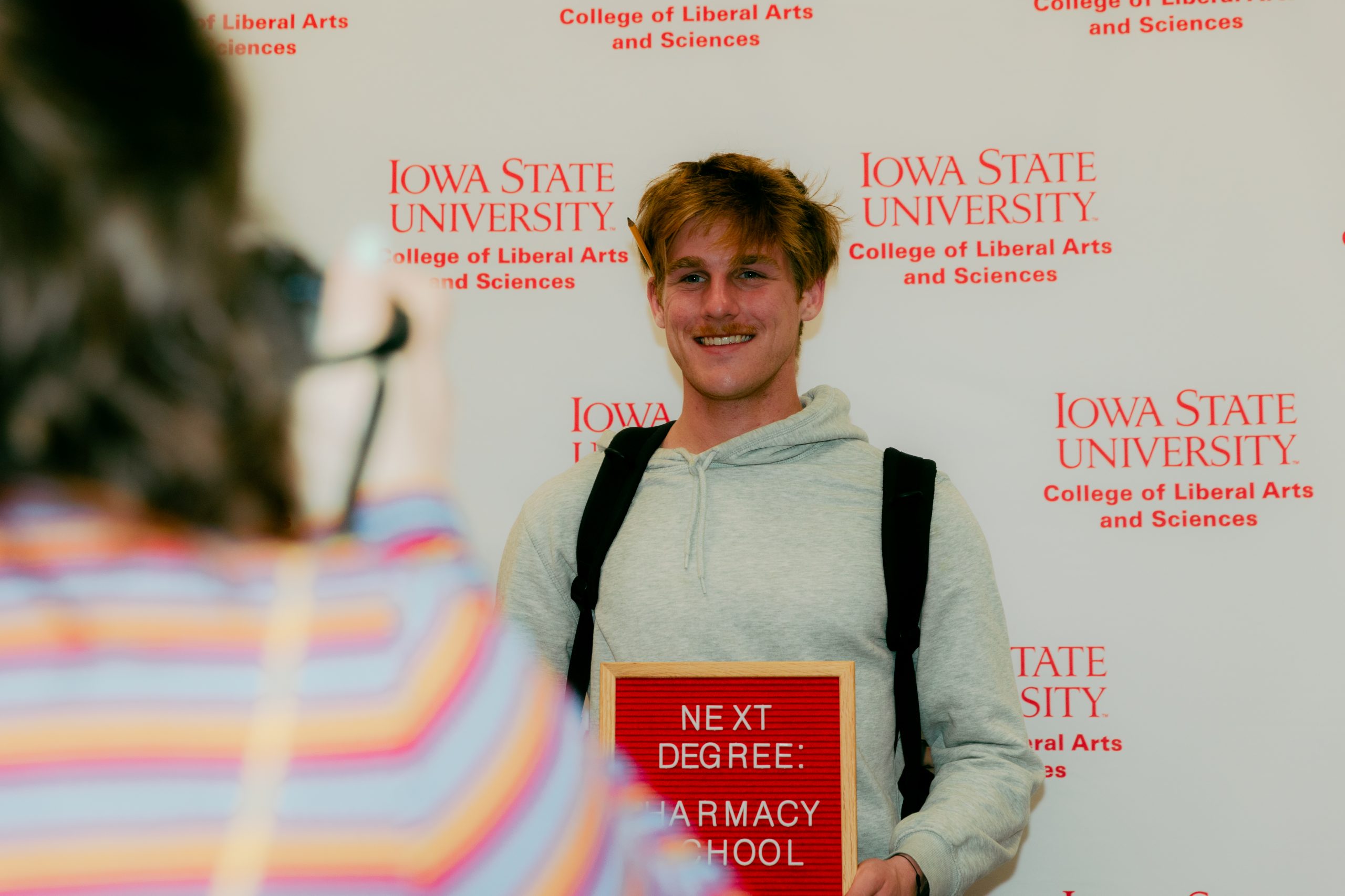 A student in a gray hoodie poses in front of an Iowa State University College of Liberal Arts and Sciences backdrop, smiling while holding a red letterboard that says, “NEXT DEGREE: PHARMACY SCHOOL.” A person in a striped shirt is seen in the foreground taking the photo.