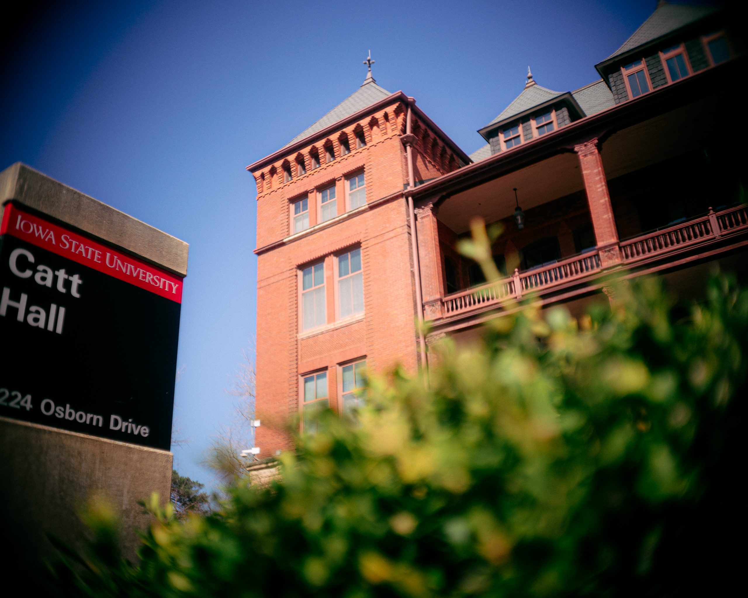 A low-angle view of Catt Hall at Iowa State University on a clear spring day, with bright green bushes in the foreground and the red-brick building with turreted rooflines and a covered balcony rising against a blue sky. A campus sign reading 'Iowa State University, Catt Hall, 2224 Osborn Drive' is partially visible on the left.