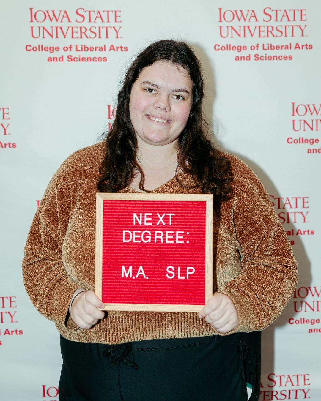 A student smiles while holding a red letter board that reads "NEXT DEGREE: M.A. SLP" in white letters. She is standing in front of a step-and-repeat banner with the Iowa State University College of Liberal Arts and Sciences logo repeated in red text. She is wearing a brown textured sweater and black pants.