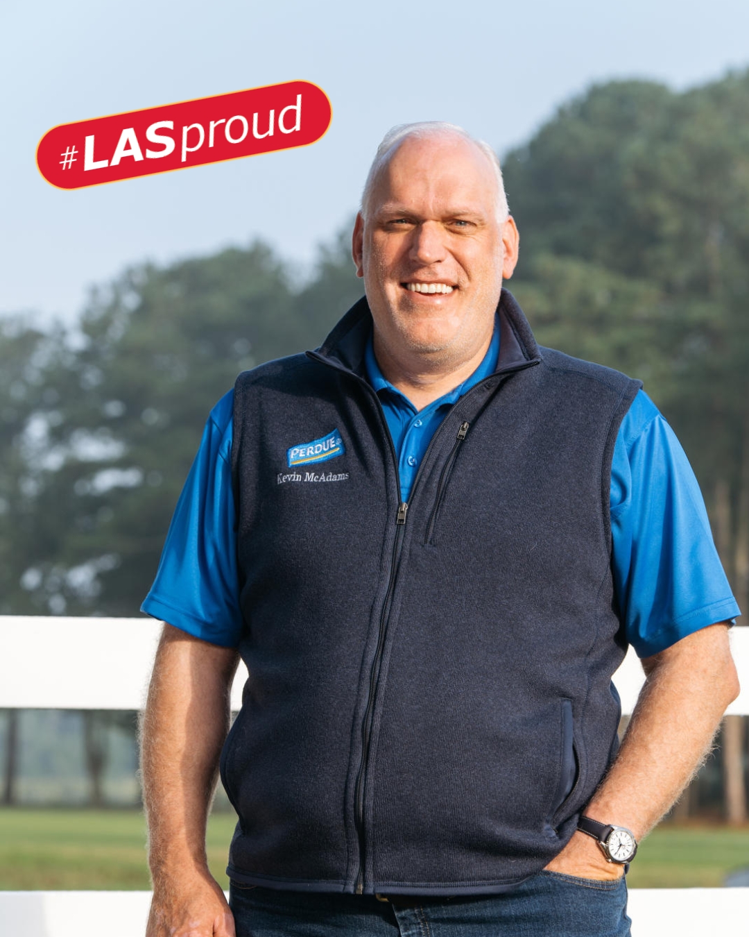A smiling man stands outdoors in front of a white fence and trees, wearing a blue polo shirt and a navy fleece vest embroidered with the Perdue Farms logo and the name 'Kevin McAdams.' A red graphic in the top left corner reads '#LASproud.'