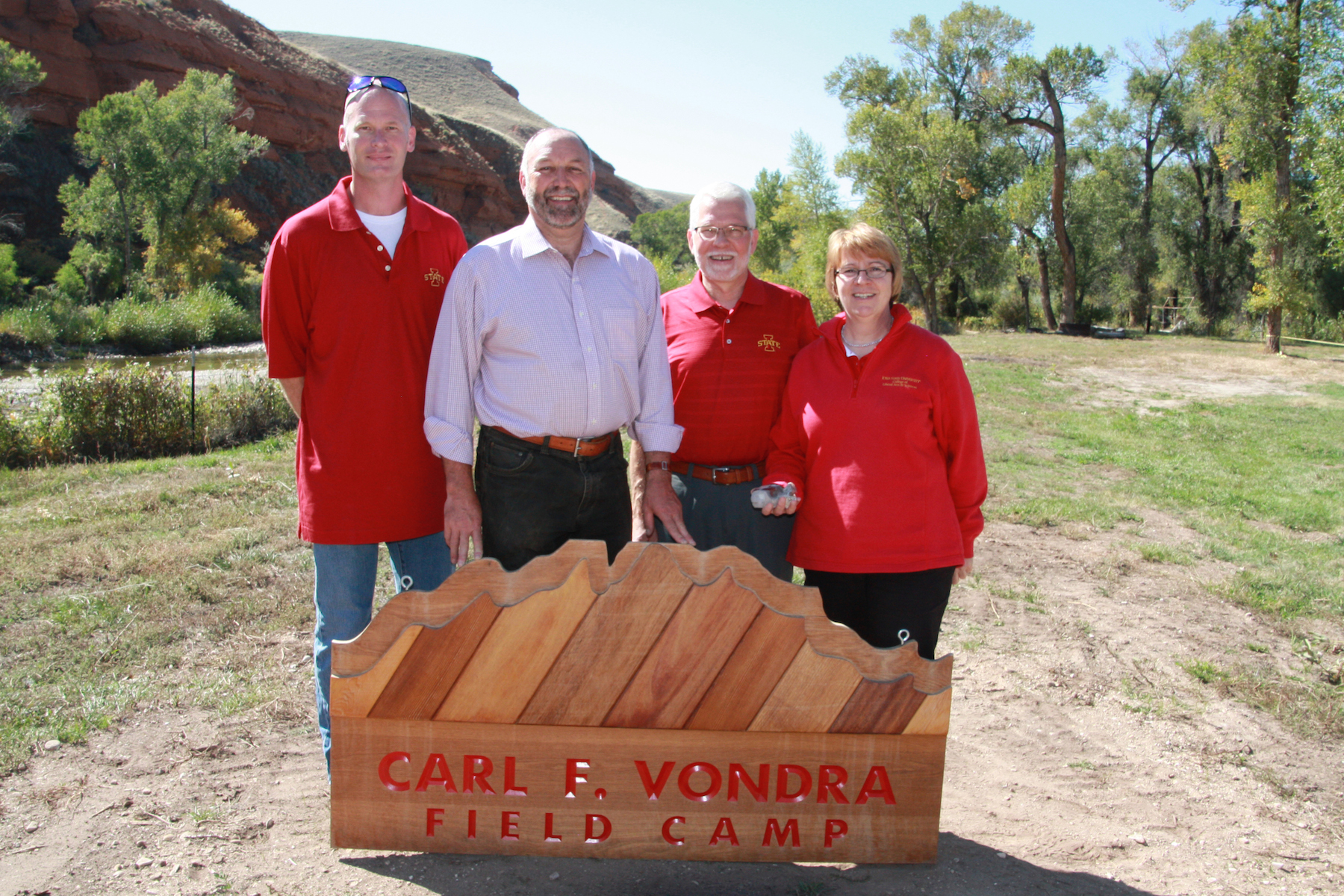 Mark Mathison, President Steven Leath, Department Chair Bill Simpkins, and LAS Dean Beate Schmittmann stand outside in front of a sign for Carl F. Vondra Field Camp. Trees and rocky cliffs are behind them.