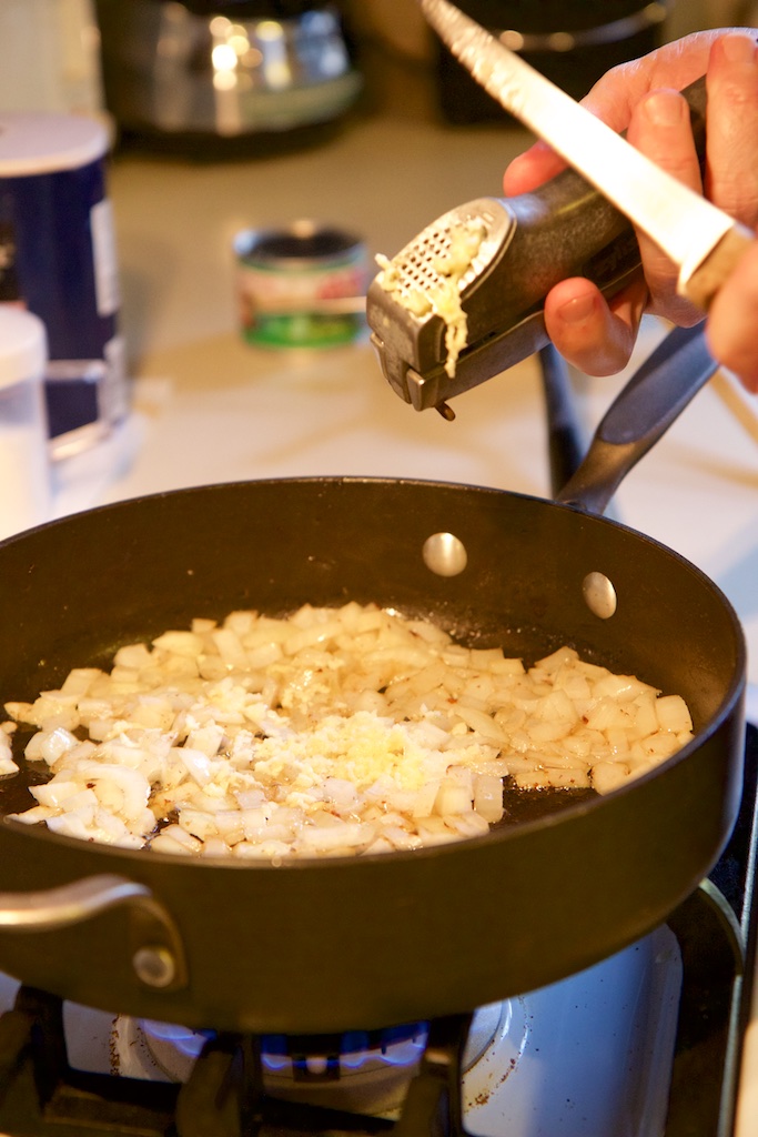 A hand holds a garlic press and a knife to scrape off the garlic into a frying pan on the stove.