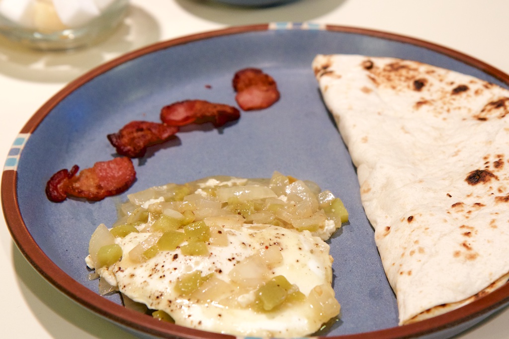 On a blue plate lies a tortilla folded in half, bits of bacon, and eggs with green chili.