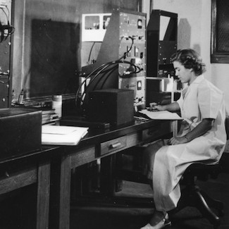 A black and white photo of Darleane Hoffman at a desk with various machines.