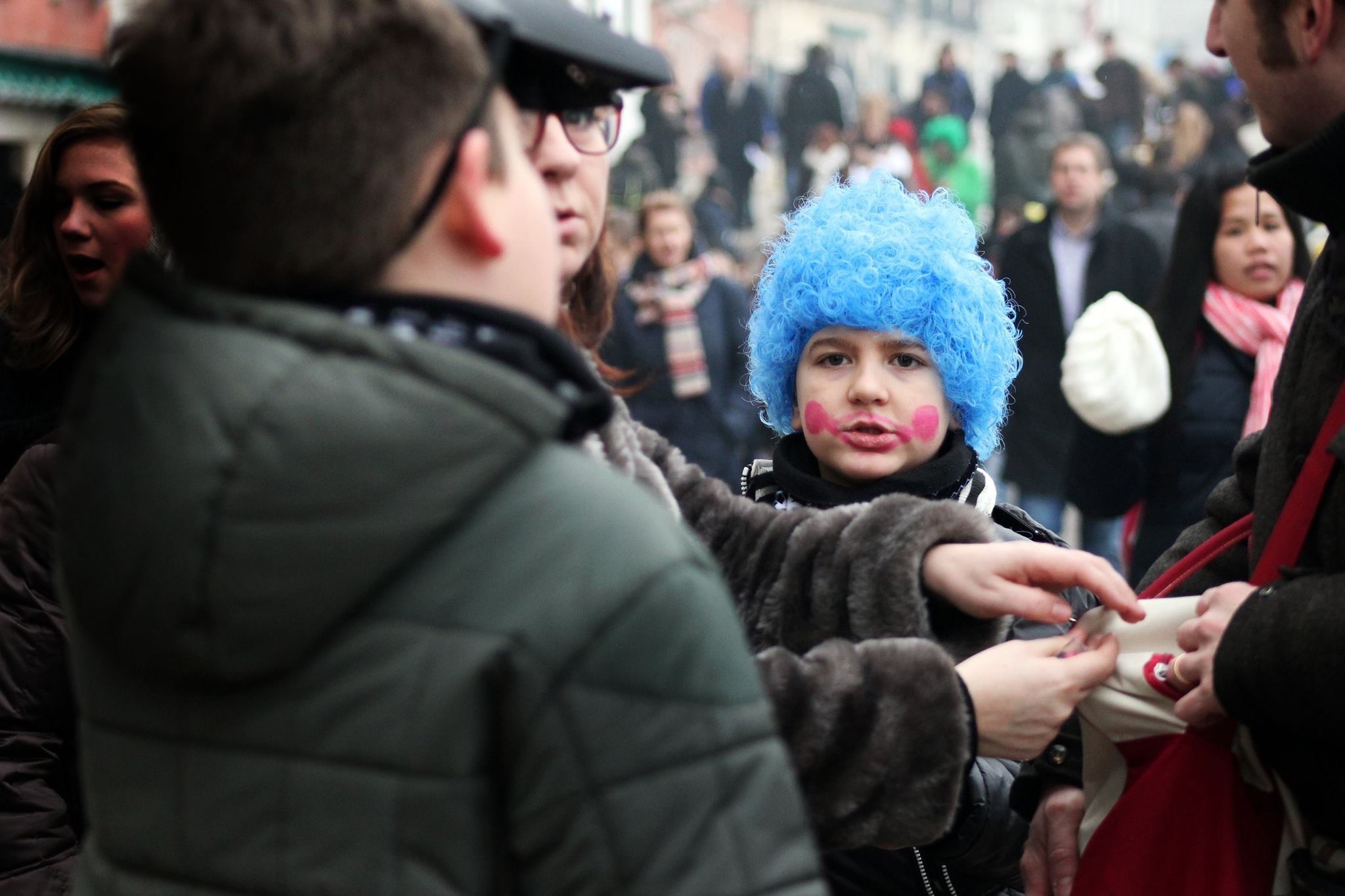 A boy wears a blue wig and has a clown-like smile painted on his face. He stands in a crowd.