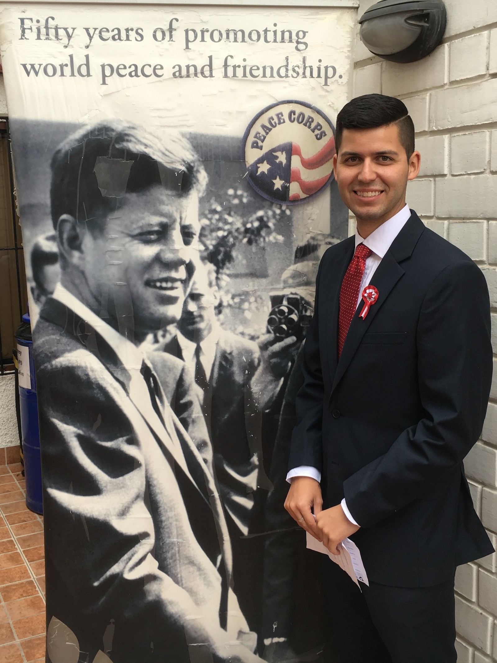 Nick Morton stands in front of a Peace Corps banner illustrated with a photo of President John F. Kennedy.