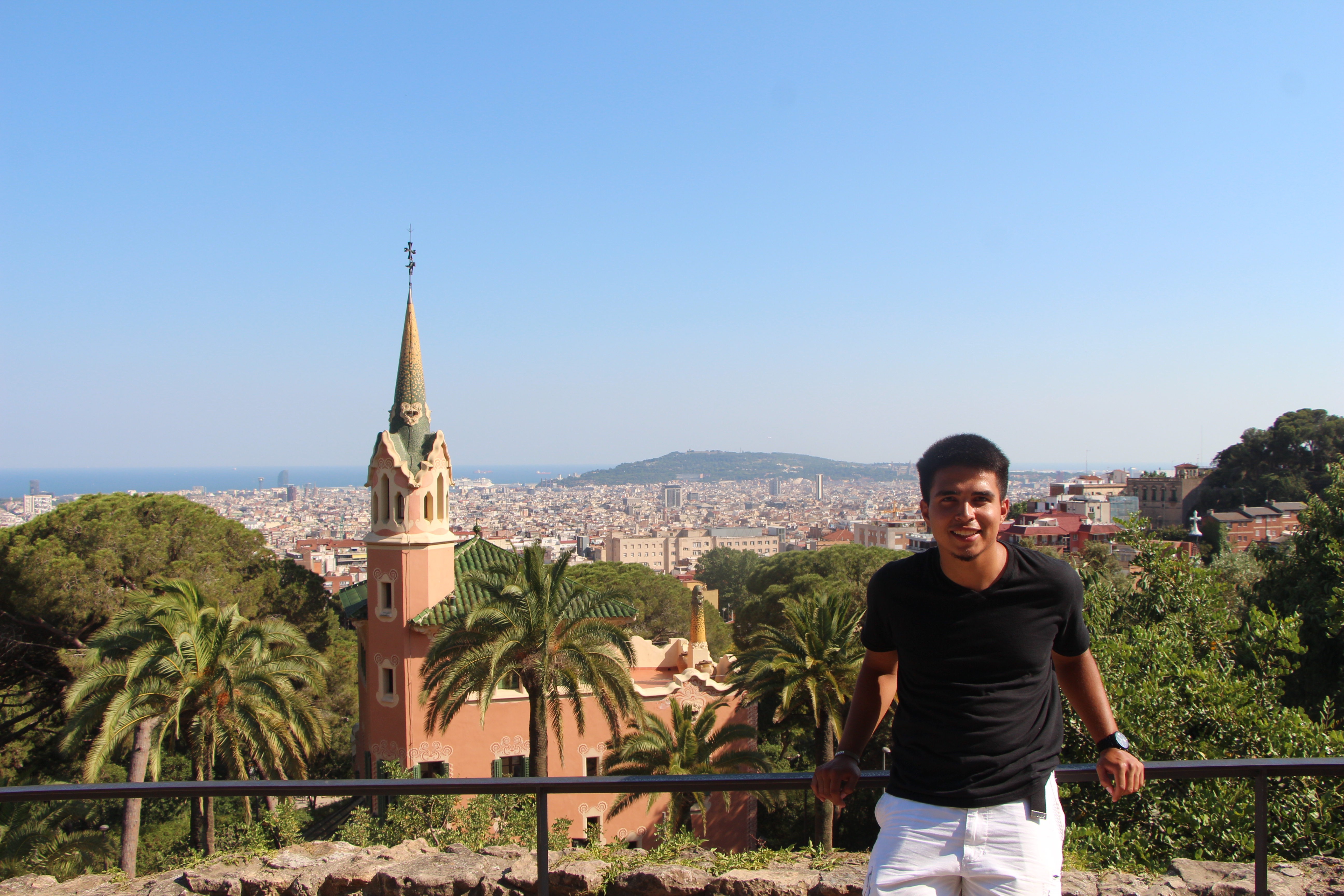 Student Rene Chavez leans against a metal railing while posing in front of a historic church in Park Guell, Barcelona, Spain. The sea and the city is in the background.