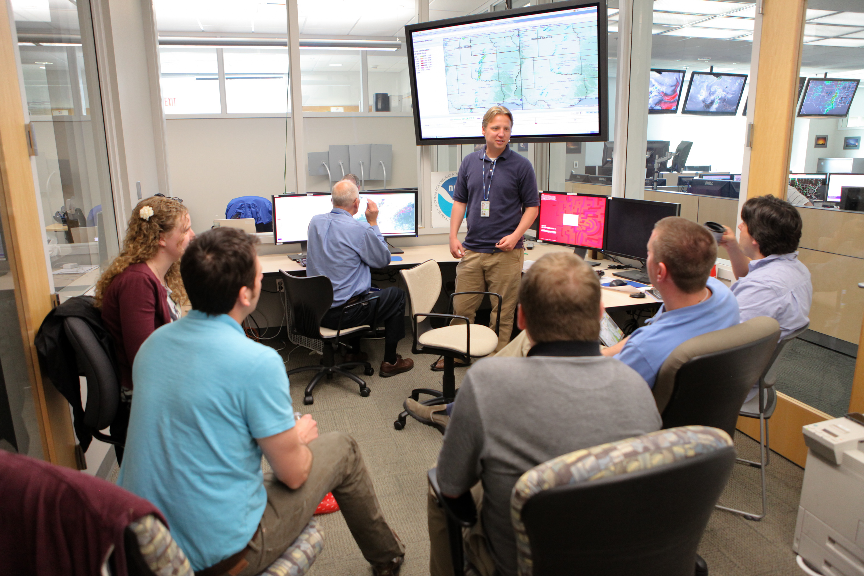 Experimenters sit in a circle. Adam Clark stands in front of monitors showing weather information.