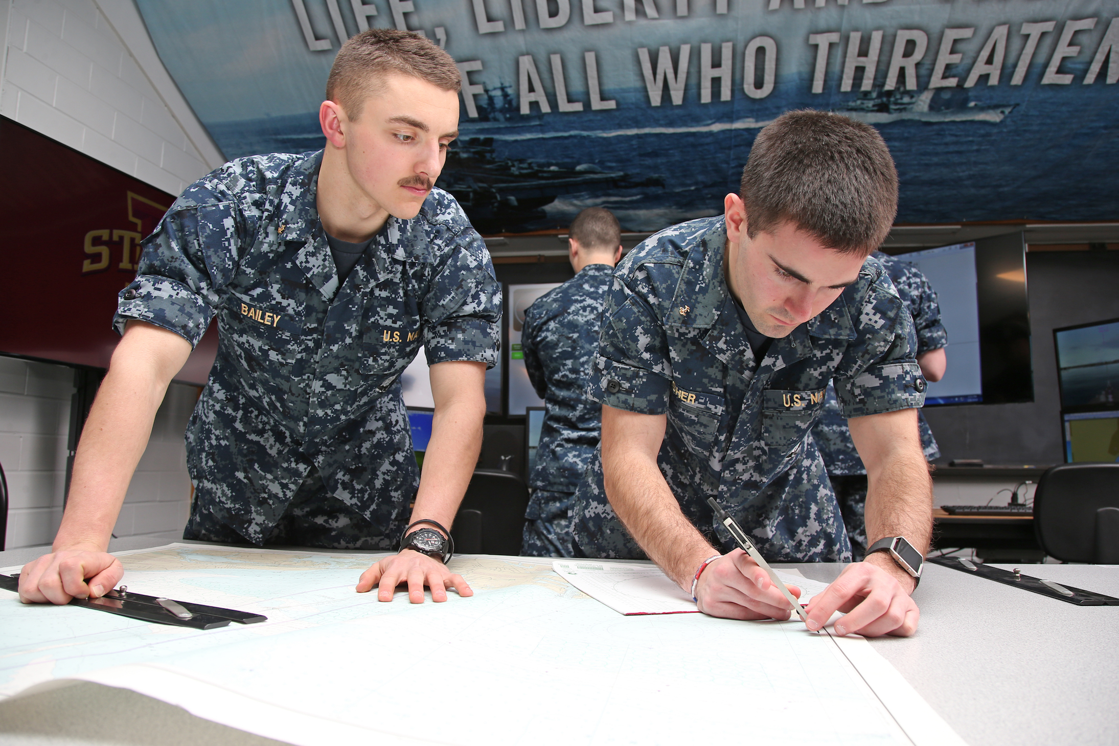 Two Navy ROTC midshipmen practice navigation skills using charts on a table.