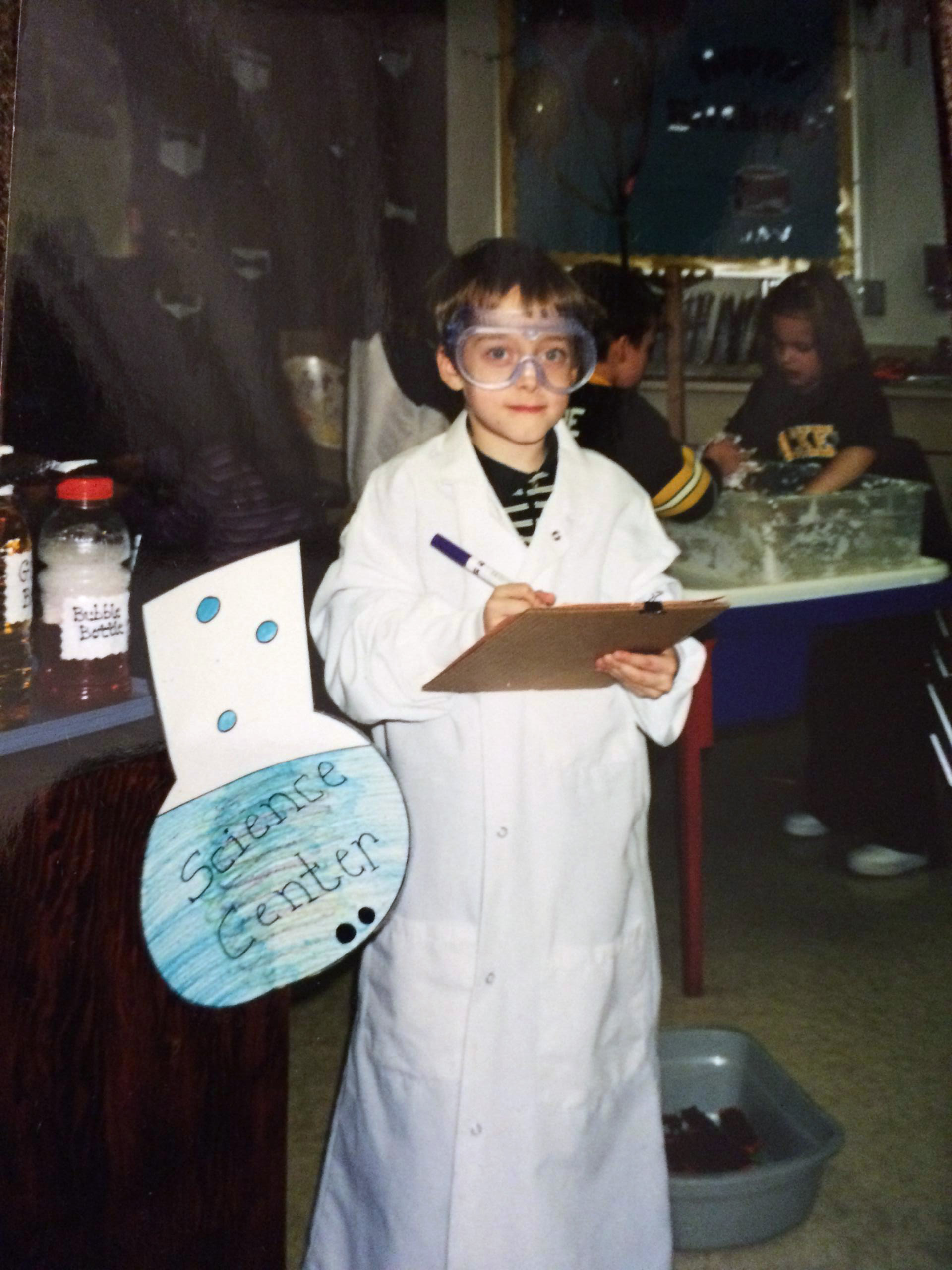 Elementary school student in lab coat and protective goggles, holding clipboard.