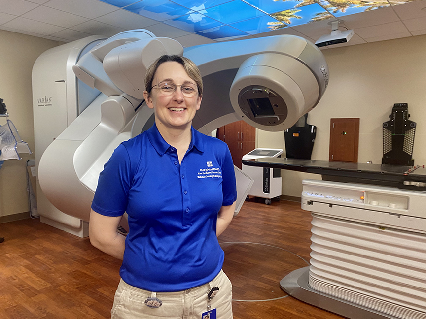 Jen Scharff poses in front of a large linear accelerator that is used to treat cancer patients