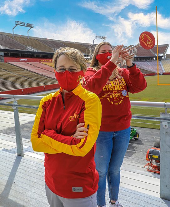 A sweeping view of Jack Trice Stadium, where Jen Scharff poses in front of Iowa State student Rebecca Gomez, who is playing her trumpet on a beautiful, sunny day