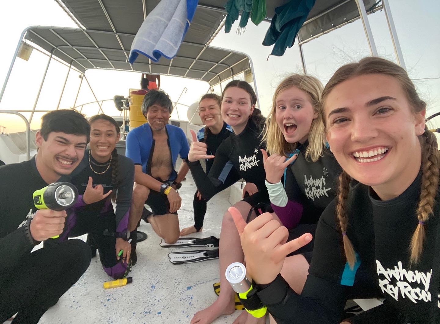 A group of students and faculty in black and blue dive suits smile at the camera while kneeling on a boat.