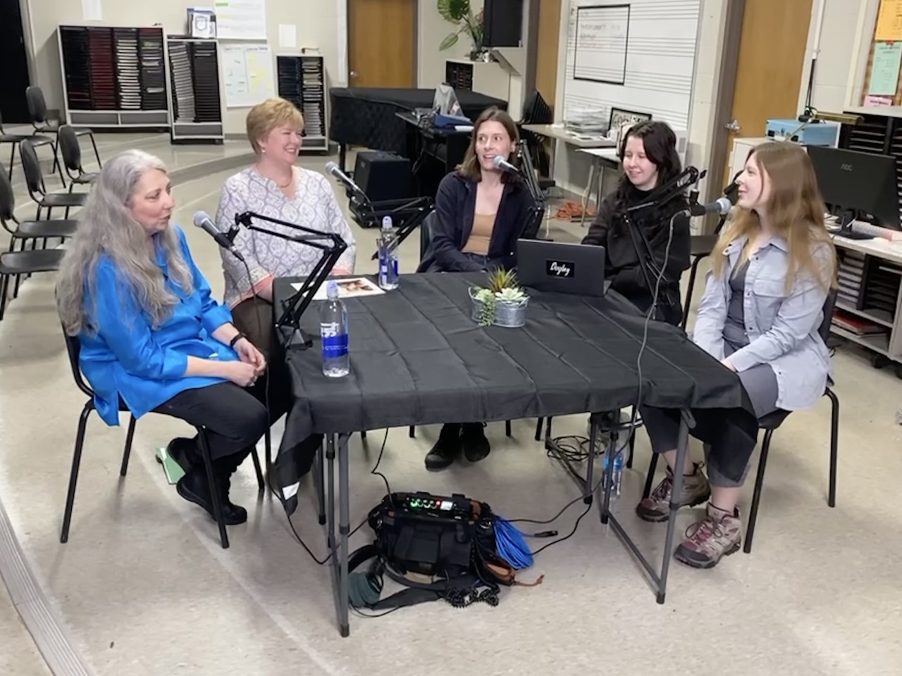 In a high school band room, five people sit around a small table covered with a black cloth. Microphones are attached to the table for each person.
